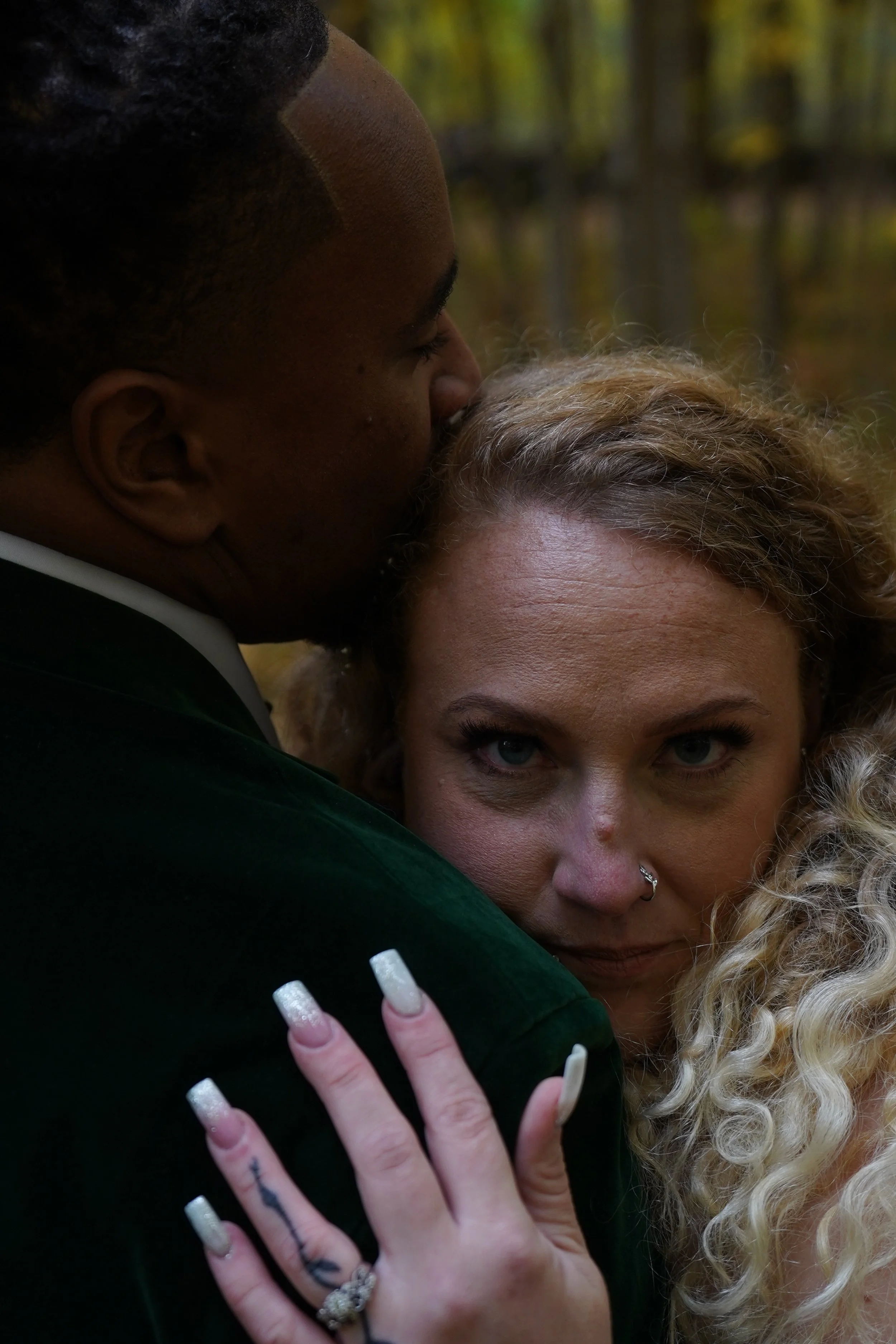A woman with curly blonde hair and a nose ring leaning against a man with dark skin and short hair, who is kissing her on the forehead, outdoors with blurred trees in the background.