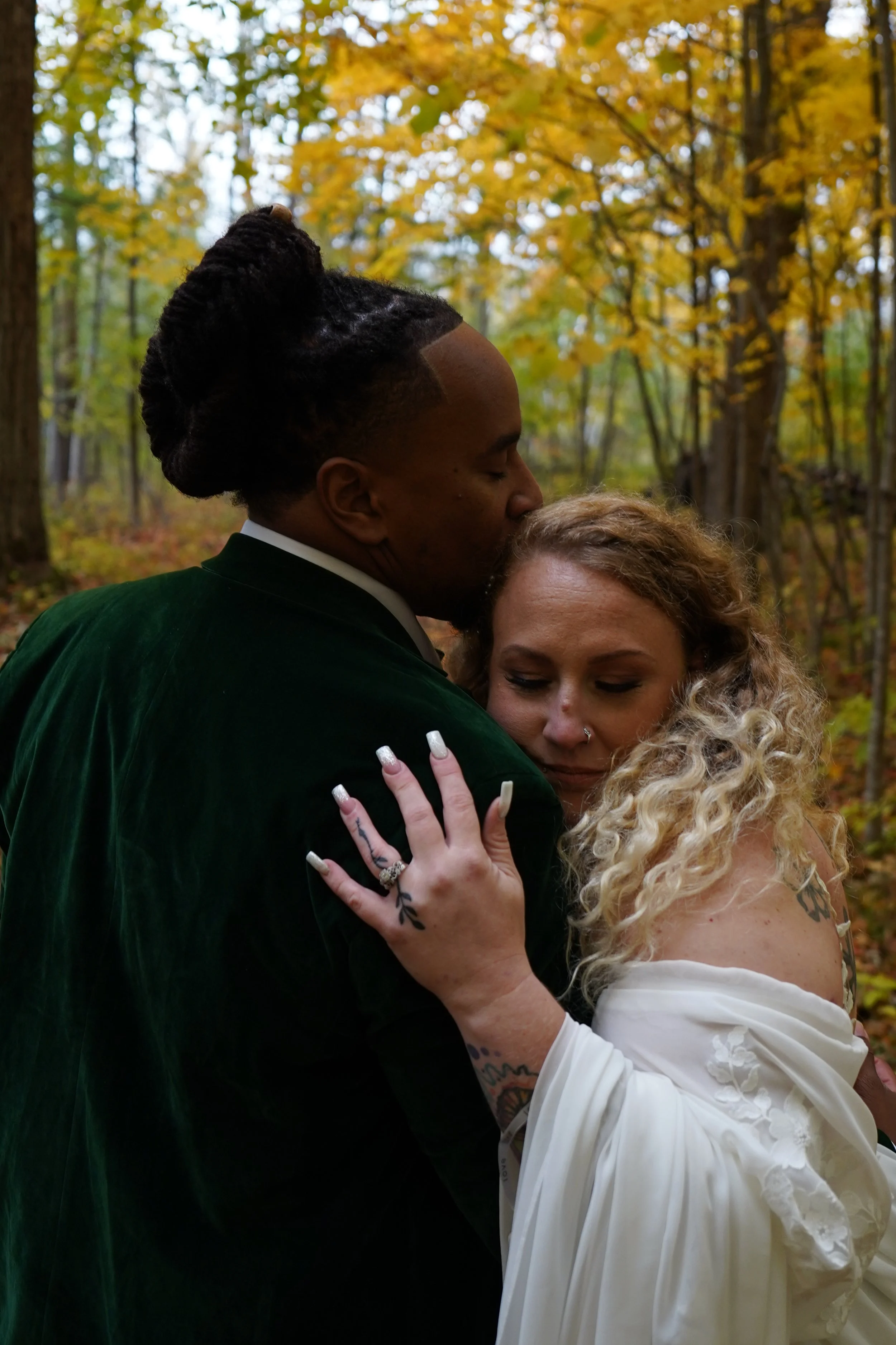 A couple sharing an intimate hug and kiss in a wooded area during autumn, with the woman resting her head on the man's chest and both dressed in formal attire.