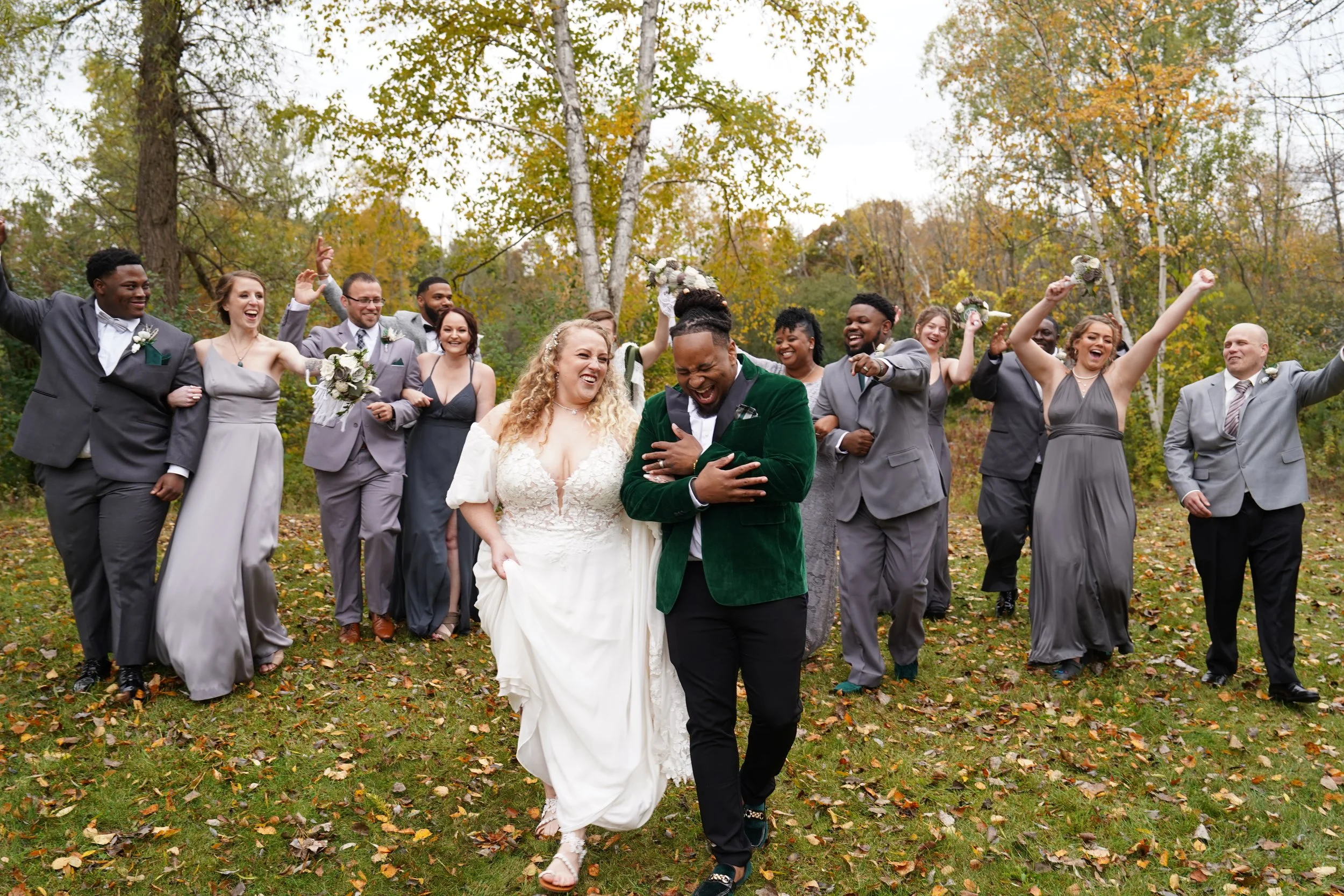 Happy wedding party walking outdoors on fall day, bride in white dress and groom in green jacket leading, bridesmaids and groomsmen in gray and silver suits following, celebrating with smiles and flower bouquets.
