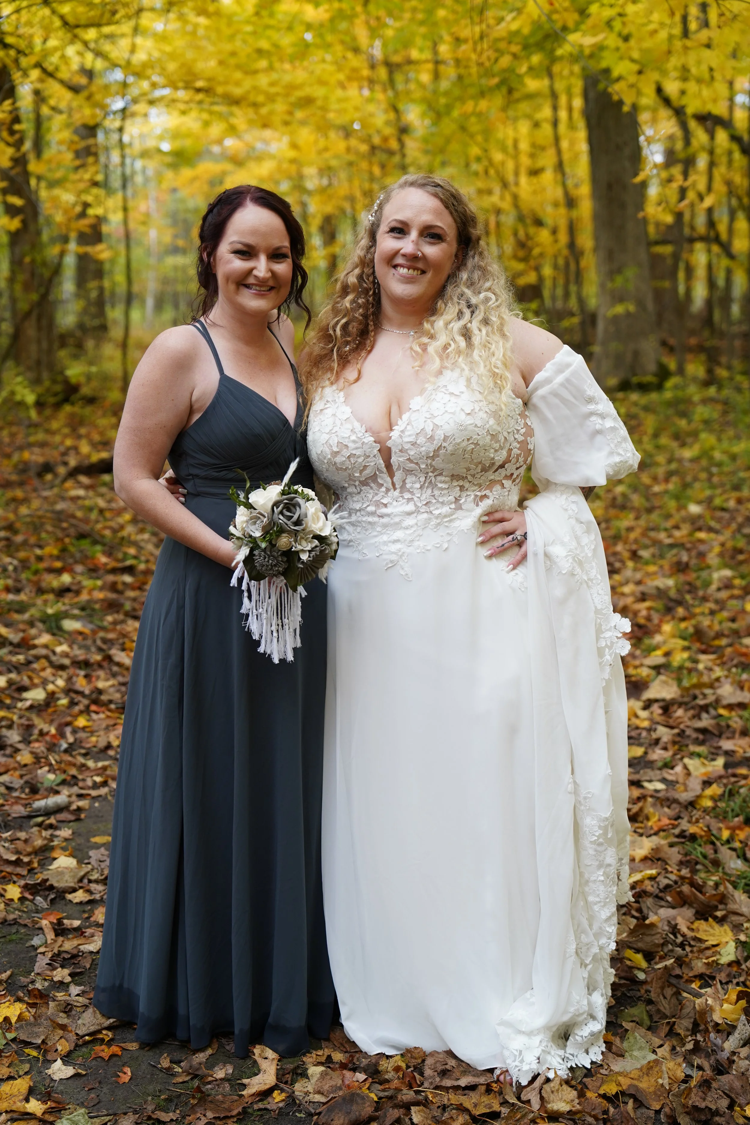 Two women, one in a white wedding dress and the other in a dark blue bridesmaid dress, standing together in a forest with autumn leaves, smiling at the camera.