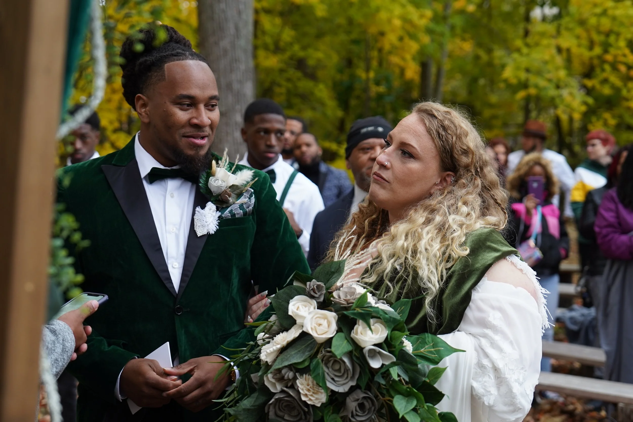 A wedding ceremony outdoors with a groom in a green velvet tuxedo and a woman with curly blond hair holding a bouquet of white roses and greenery, surrounded by guests.