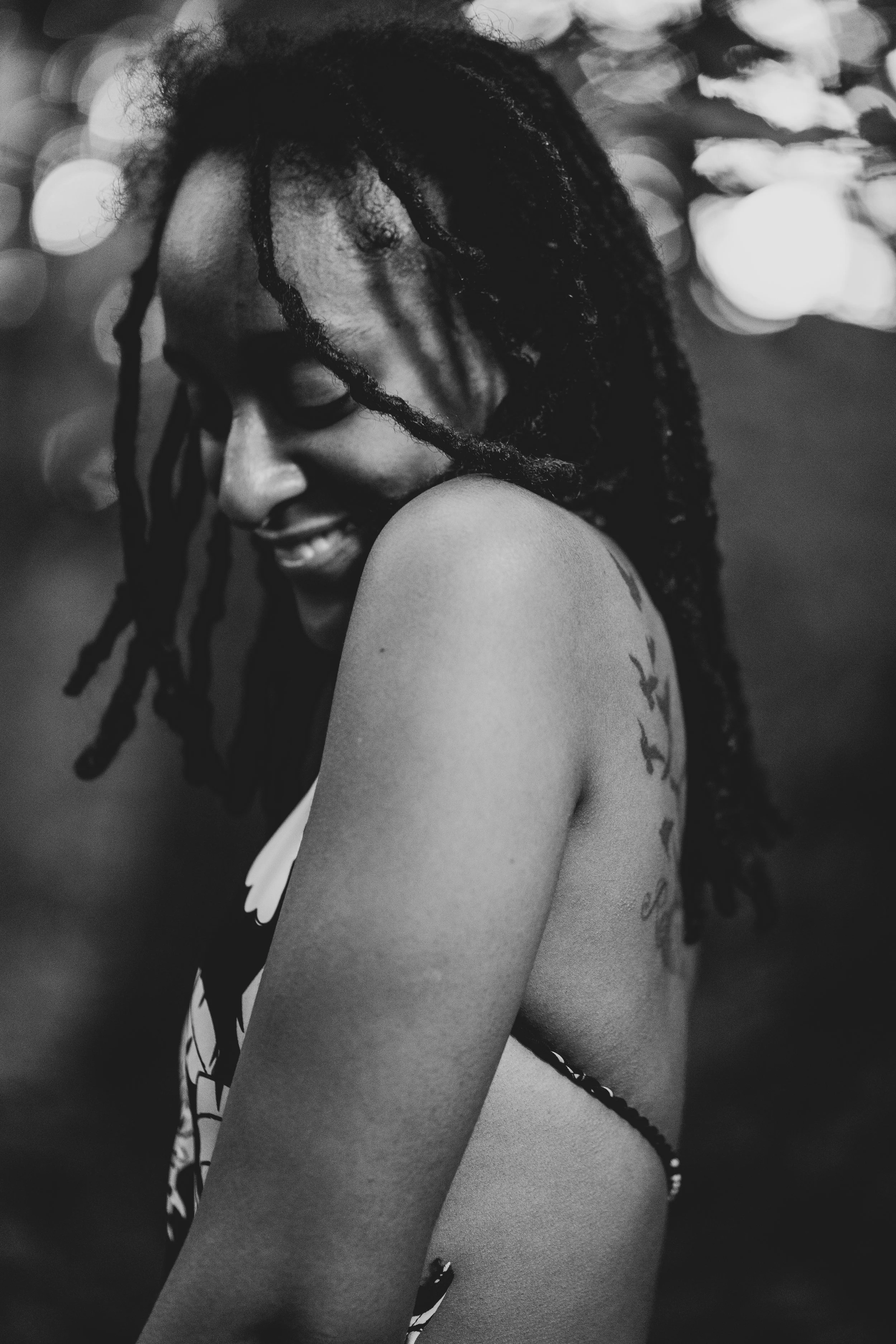 A smiling woman with dreadlocks is turned to the side with her eyes closed, showing her shoulder and tattoos on her arm and side, in a black-and-white photo with blurred bokeh lights in the background.