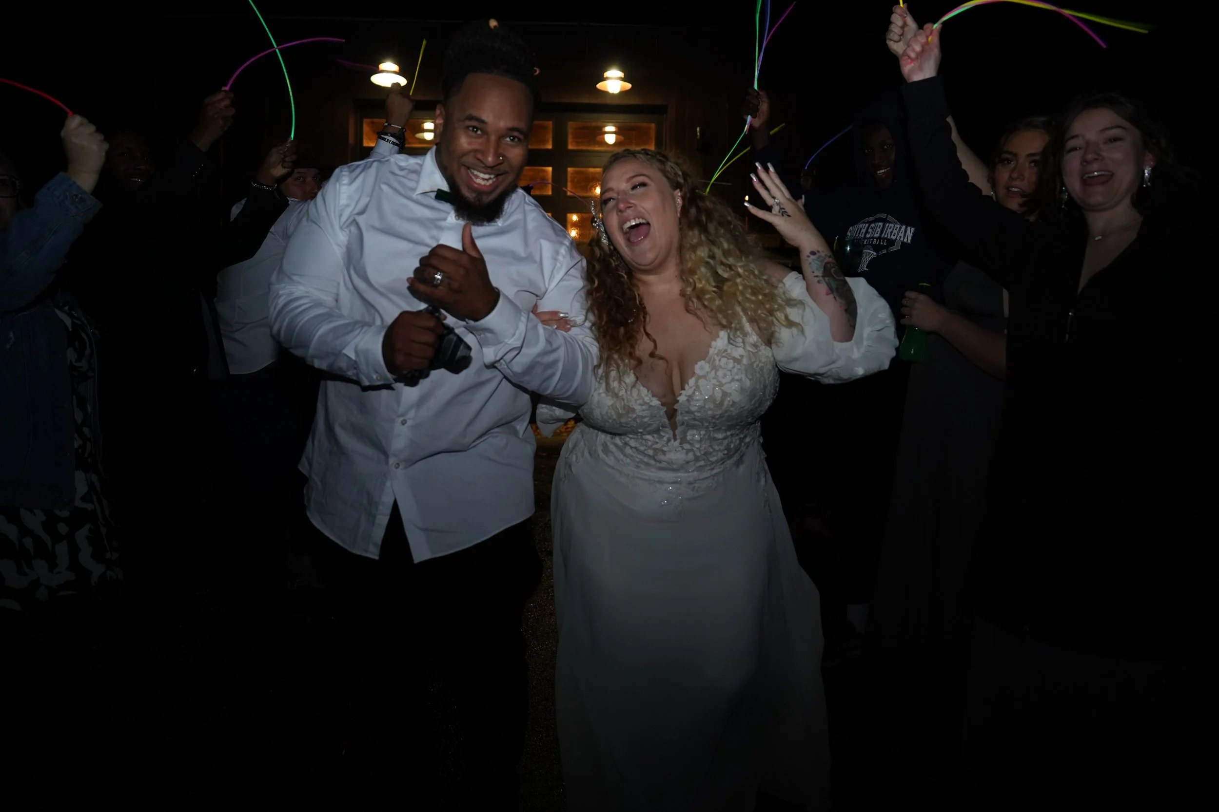 A wedding reception with a group of people celebrating, including a woman in a white wedding dress and a man in a white shirt with a camera, surrounded by others holding glow sticks and celebrating.