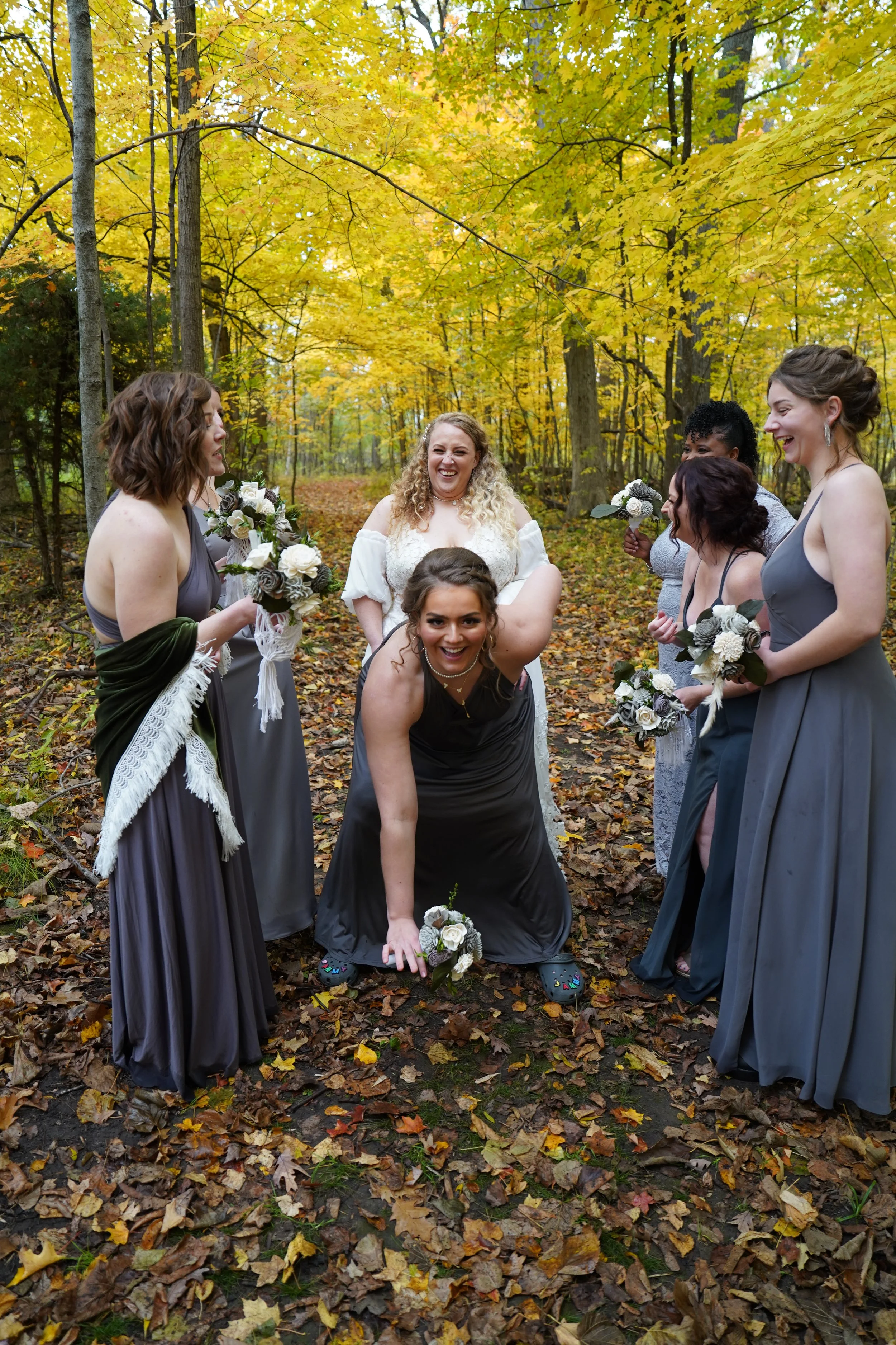A group of women, including a bride in white and bridesmaids in gray and silver dresses, laughing and posing playfully in a forest with autumn leaves on the ground and trees with yellow leaves.