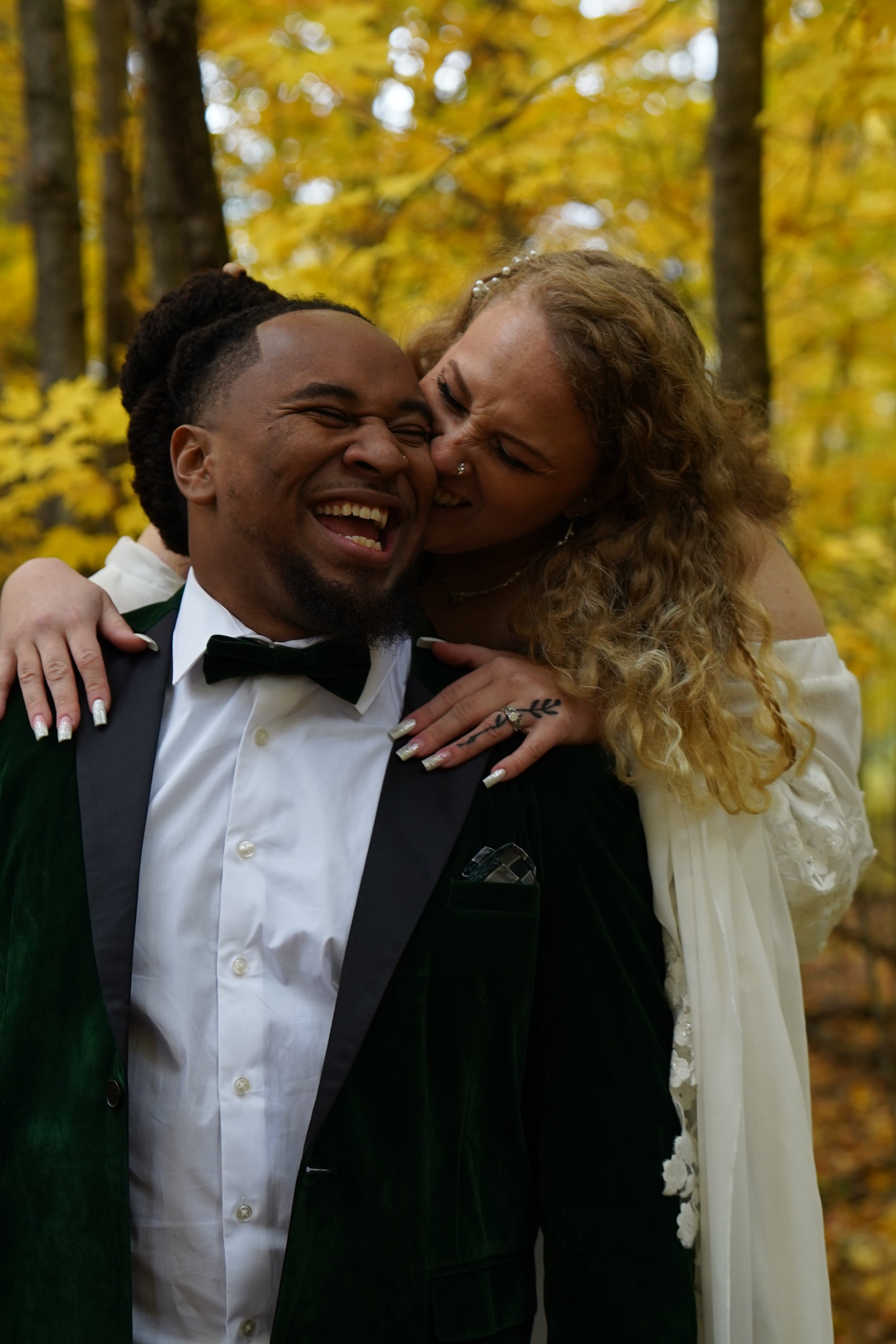 A joyful couple, the man dressed in a tuxedo with a bow tie, and the woman in a white dress, sharing a happy moment outdoors in the autumn forest, with yellow and orange leaves.