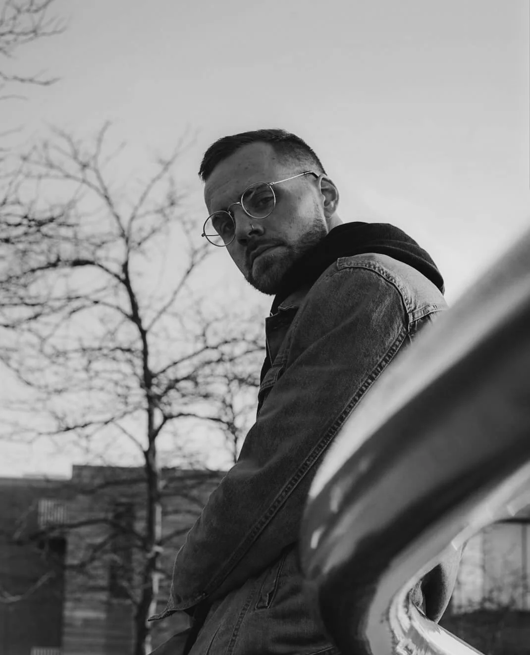 A black-and-white photo of a man with glasses leaning against a railing outdoors, with trees and buildings in the background.