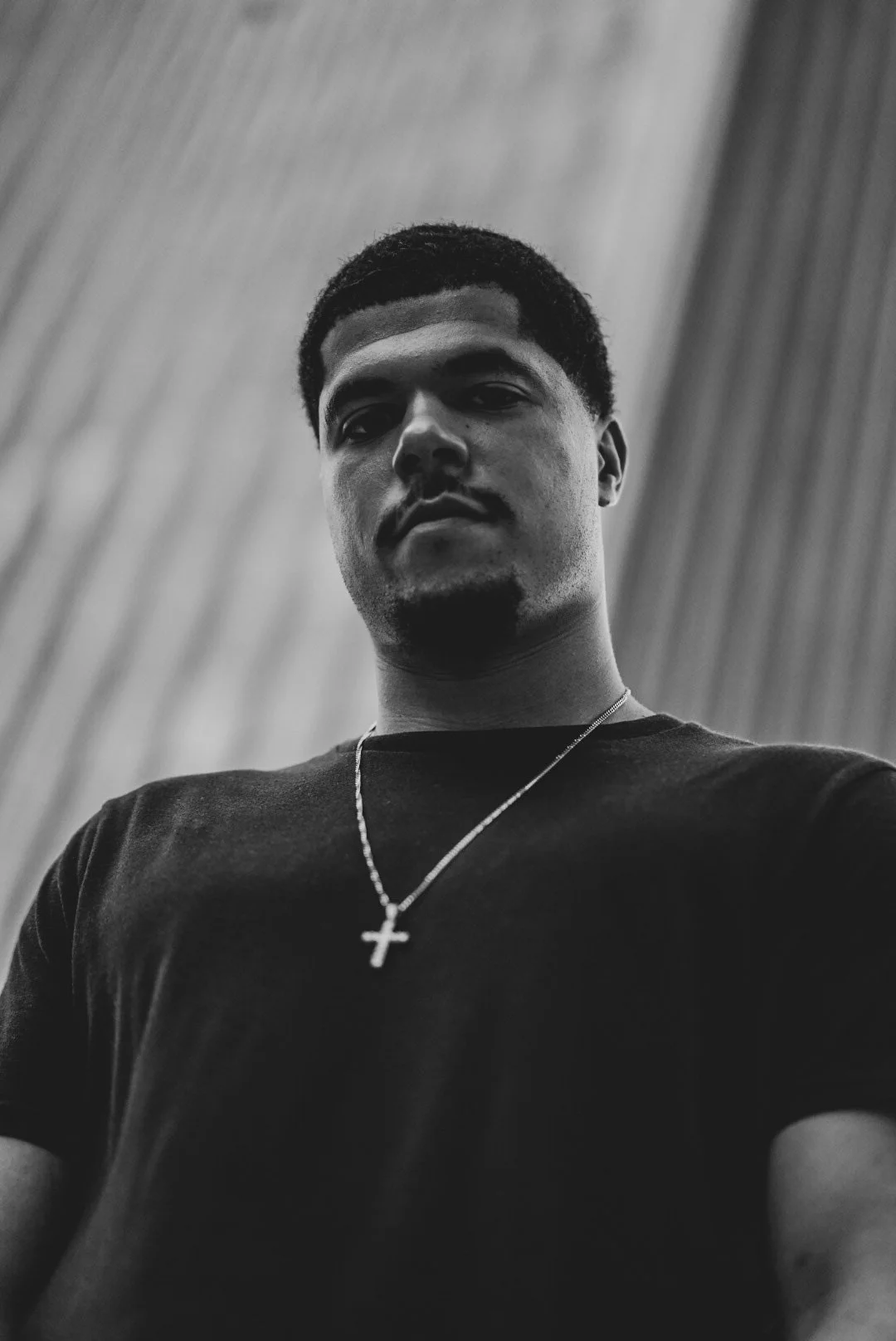 Black and white close-up photo of a young man with short hair, wearing a black shirt and a necklace with a cross pendant, looking down at the camera against a textured background.