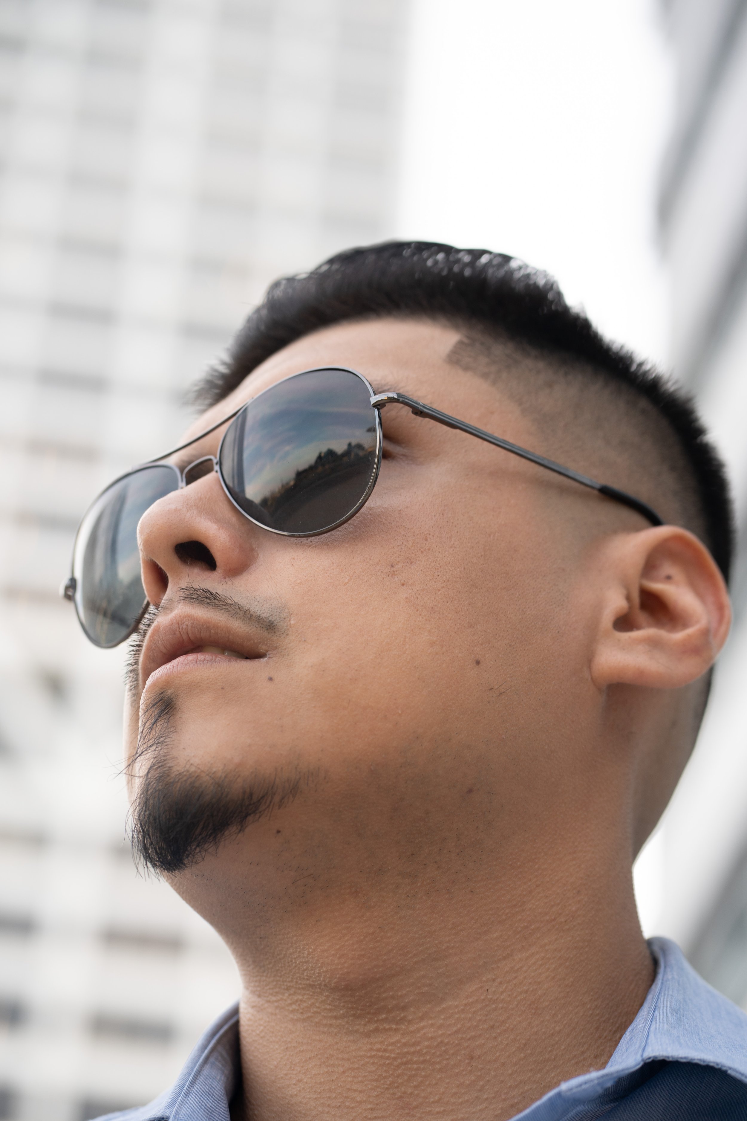 Close-up of a young man wearing sunglasses, looking upwards with a cityscape in the reflection of his sunglasses, and a modern building in the background.