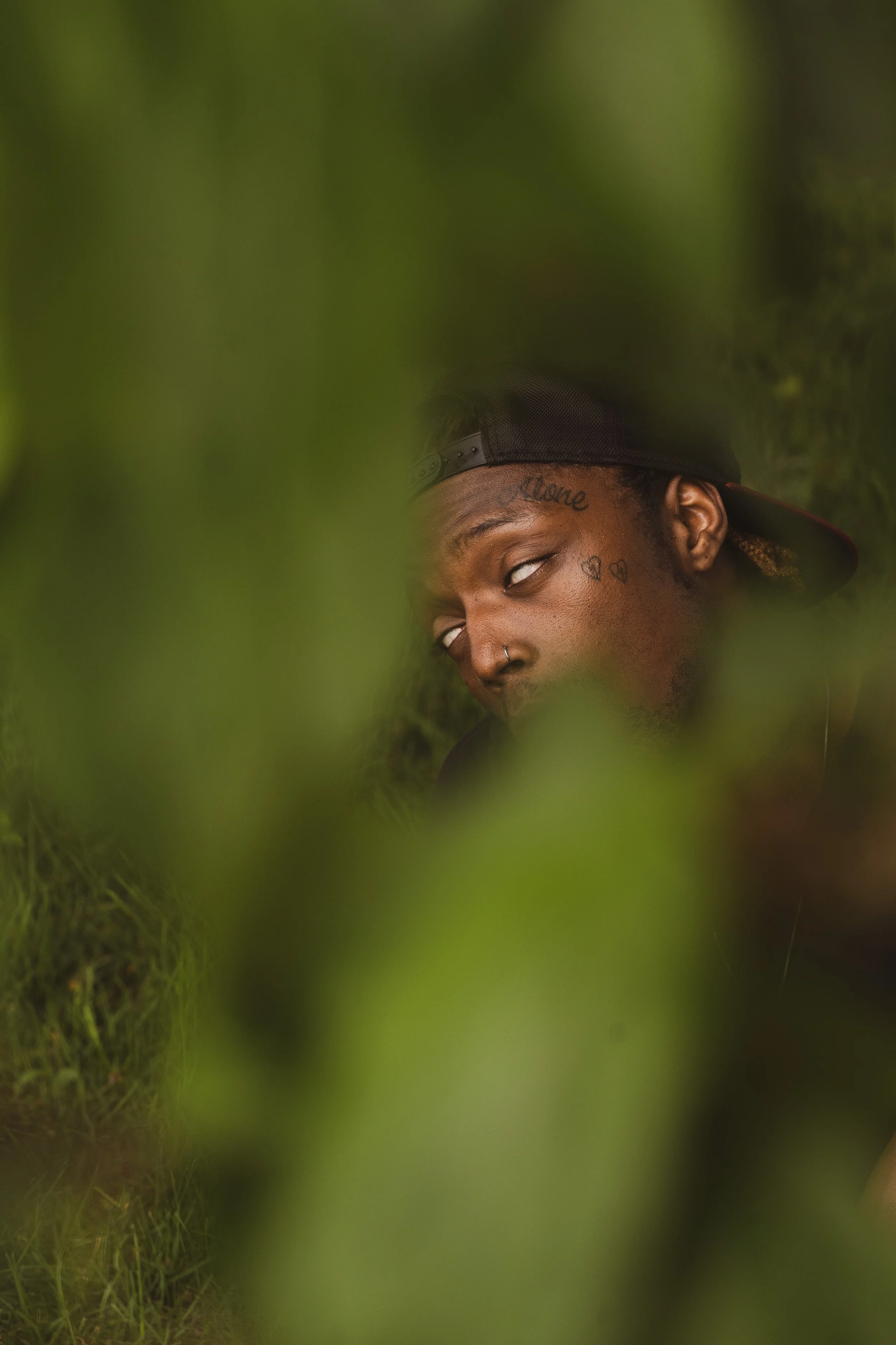 A young man with tattoos, a nose ring, wearing a baseball cap backward, looking sideways through greenery.