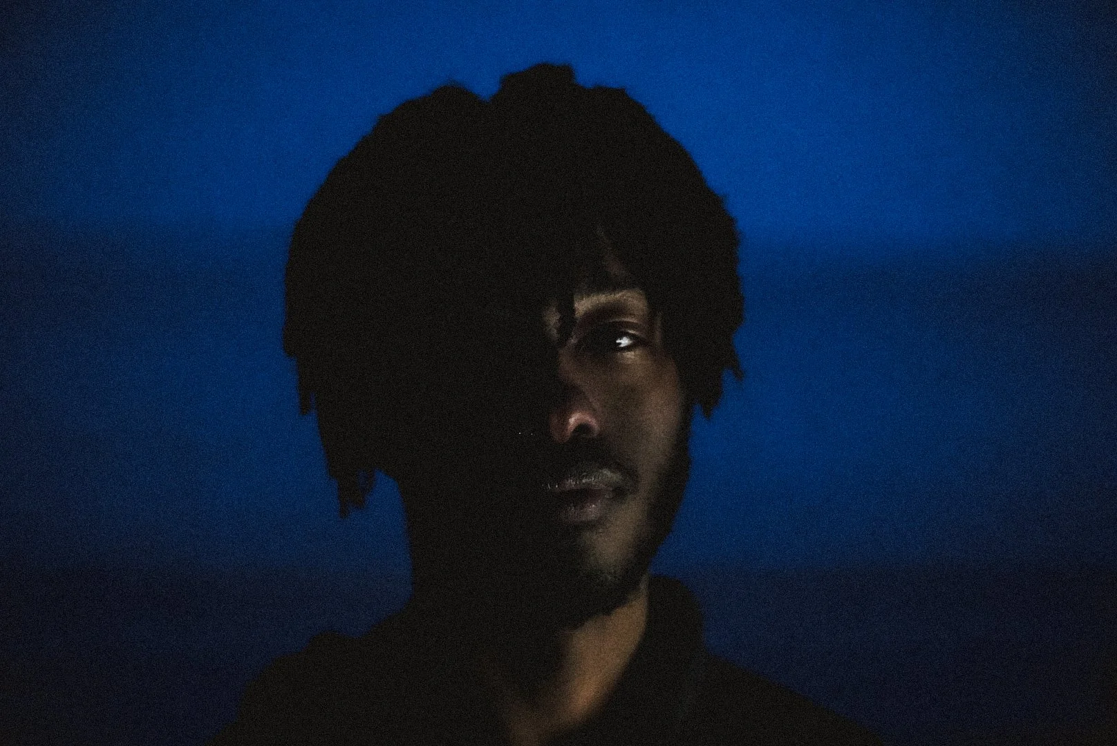 A dark portrait of a person with black hair and a beard, partially illuminated, against a blue background.