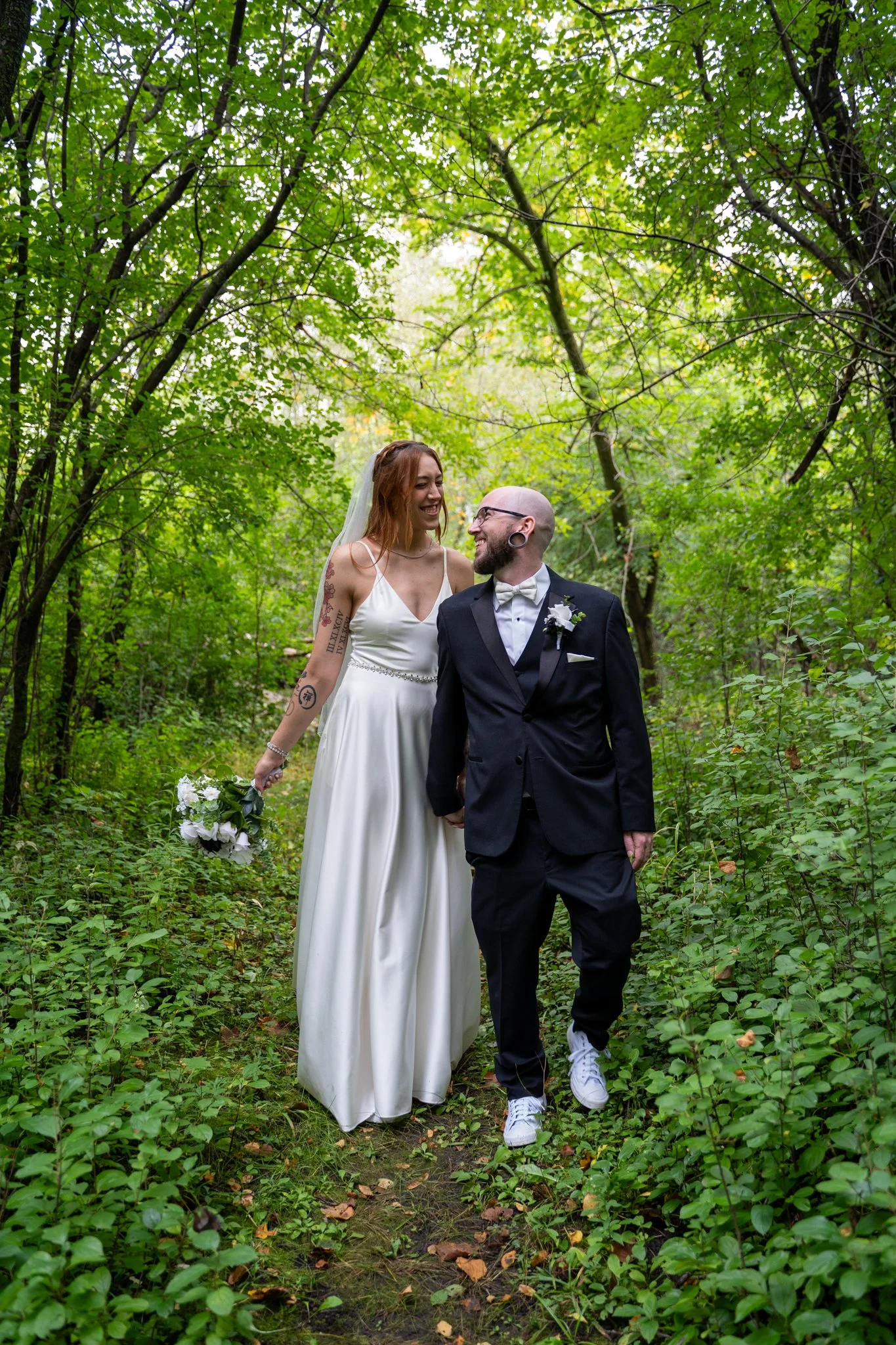 A bride and groom walking together in a lush green forest, smiling at each other, capturing a joyful moment of their wedding day.