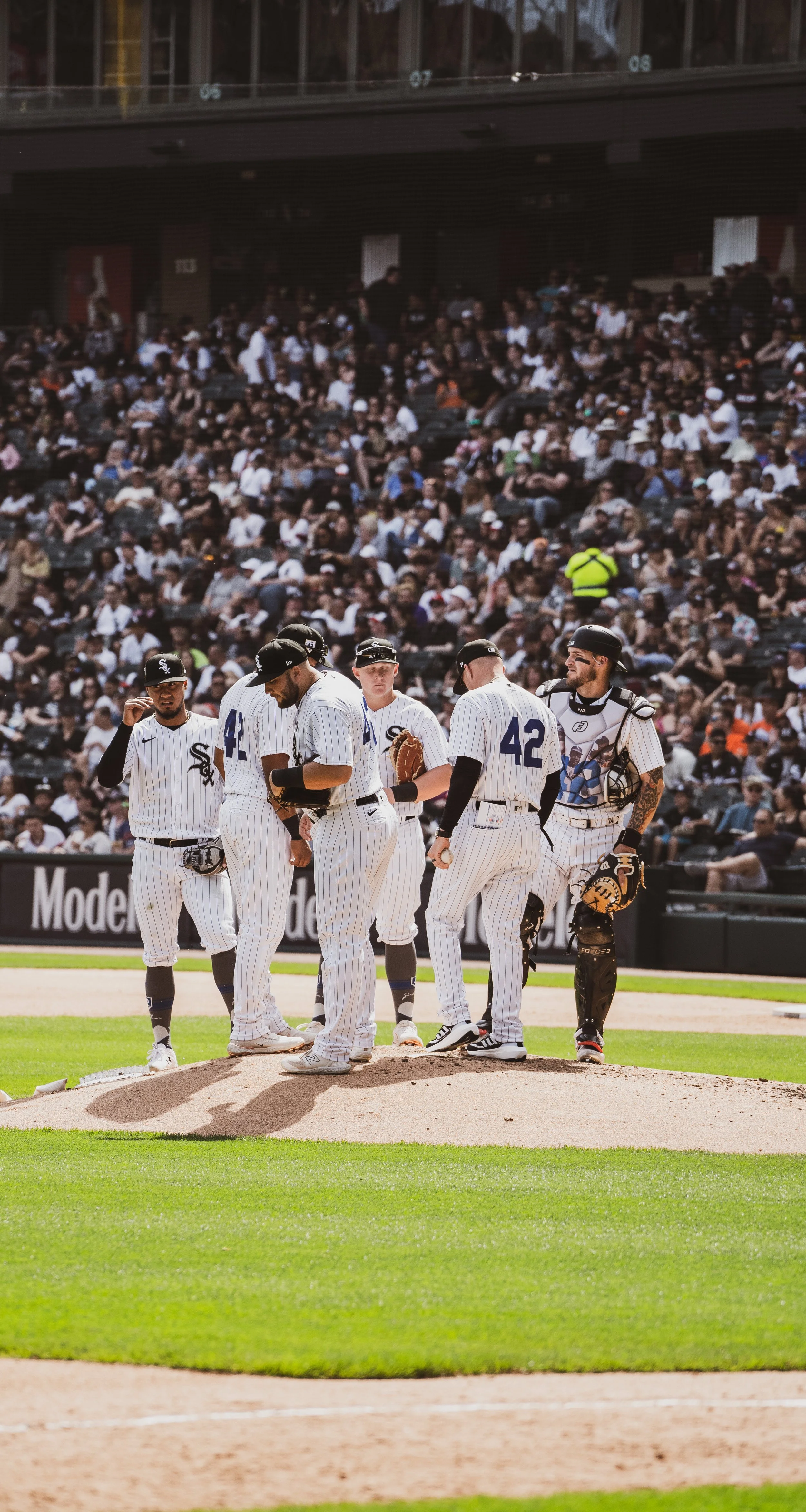 A group of baseball players in white pinstripe uniforms gathered on the mound during a game, with a large crowd in the stands behind them.