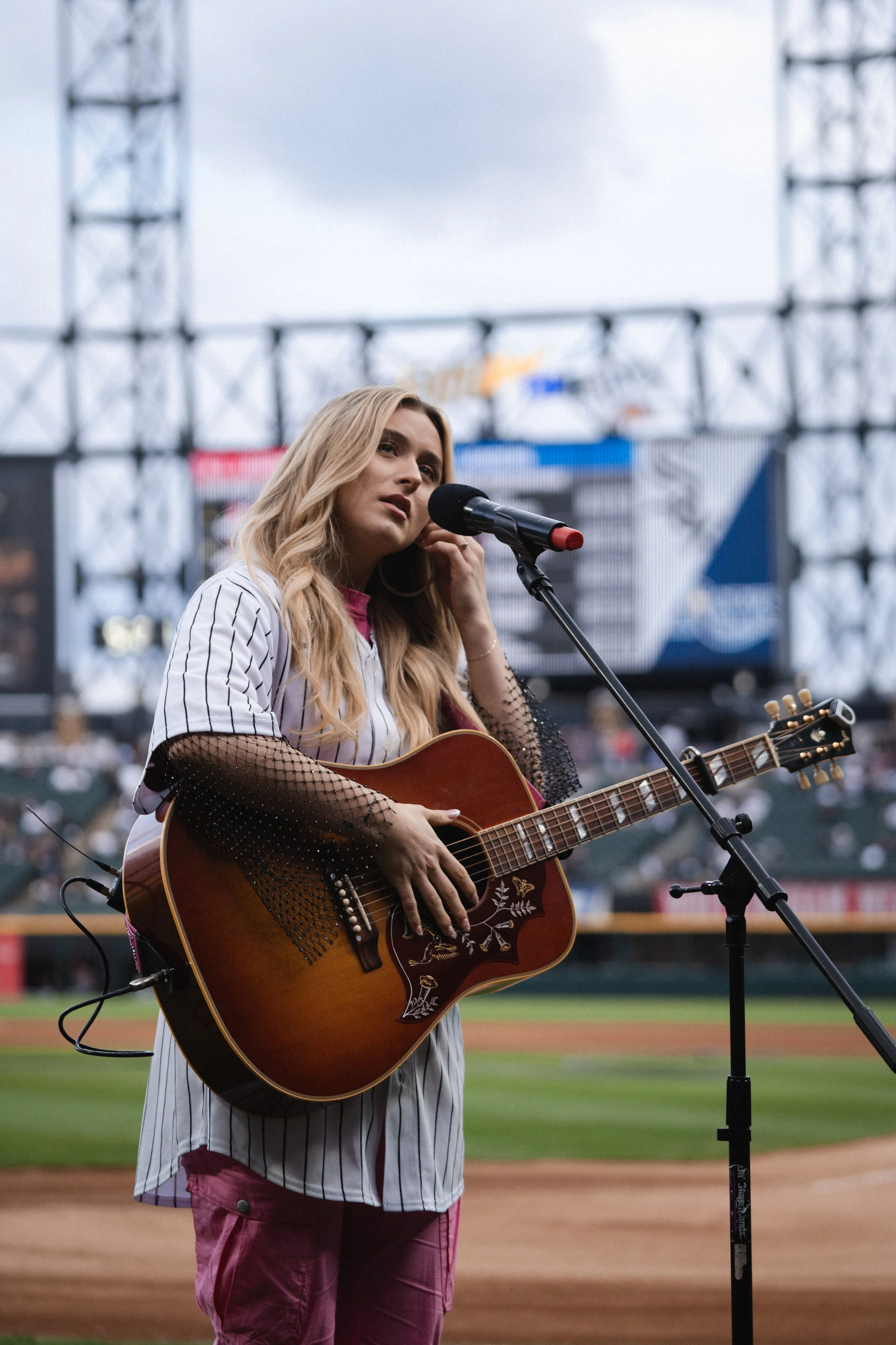 A woman with long blonde hair playing an acoustic guitar on a baseball field, with a microphone in front of her, during a concert or performance. Harper Grace