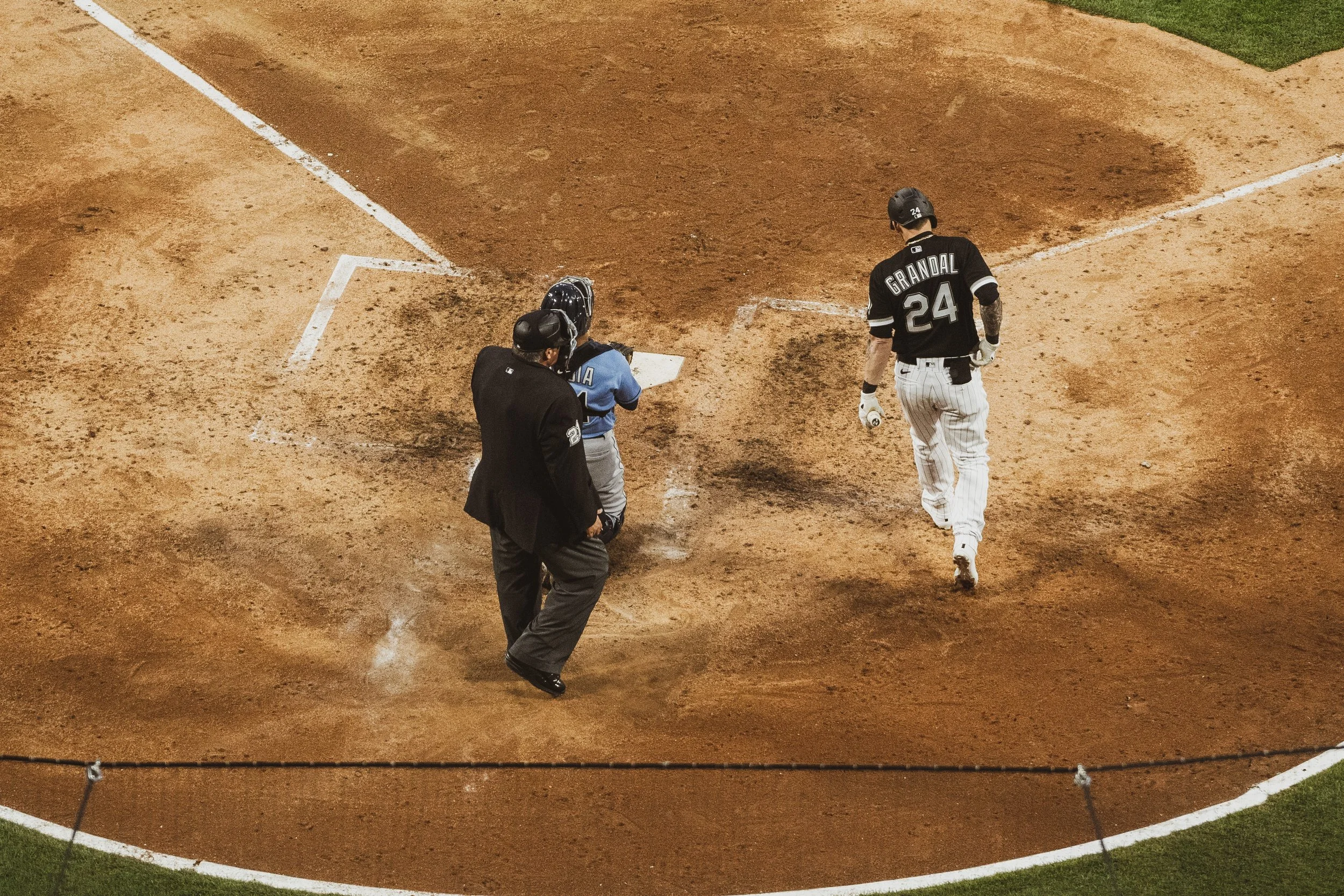 A baseball player, wearing a black jersey with the name Grandal and number 24, is running around the bases while another player in a blue jersey and a coach or umpire in black watch from nearby. The scene takes place on a baseball field.