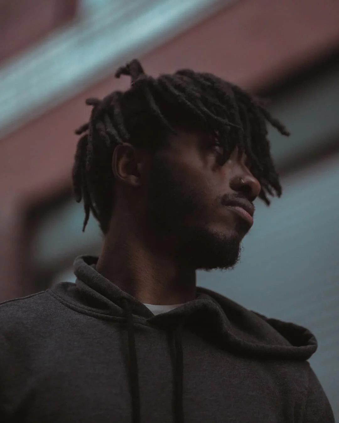 A young man with dreadlocks and a nose ring, wearing a dark hoodie, looking down thoughtfully in a dimly lit room.