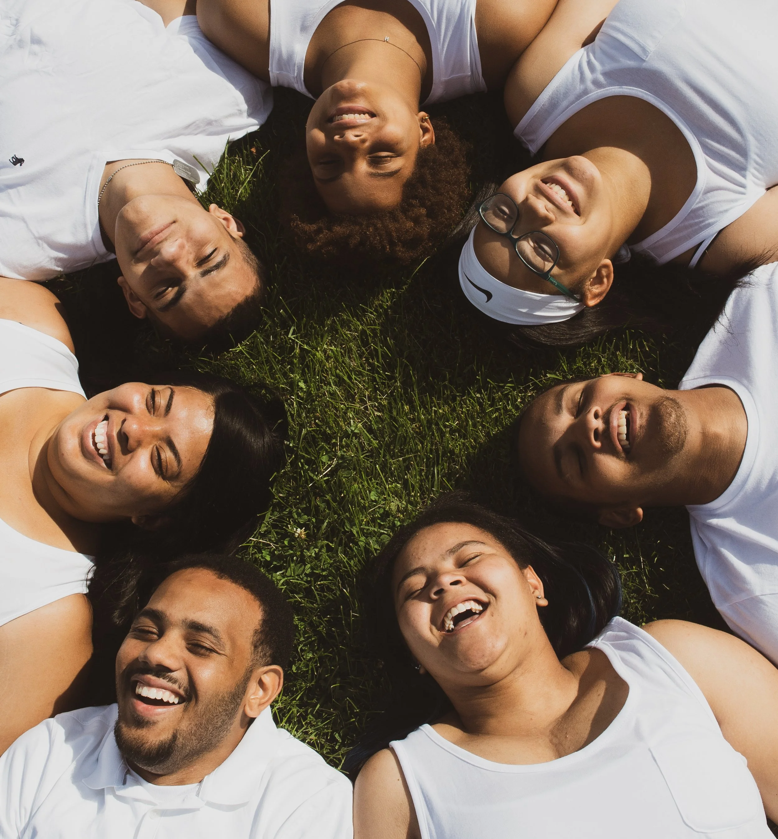 A diverse group of people lying on the grass in a circle, smiling and looking up at the camera. A Family
