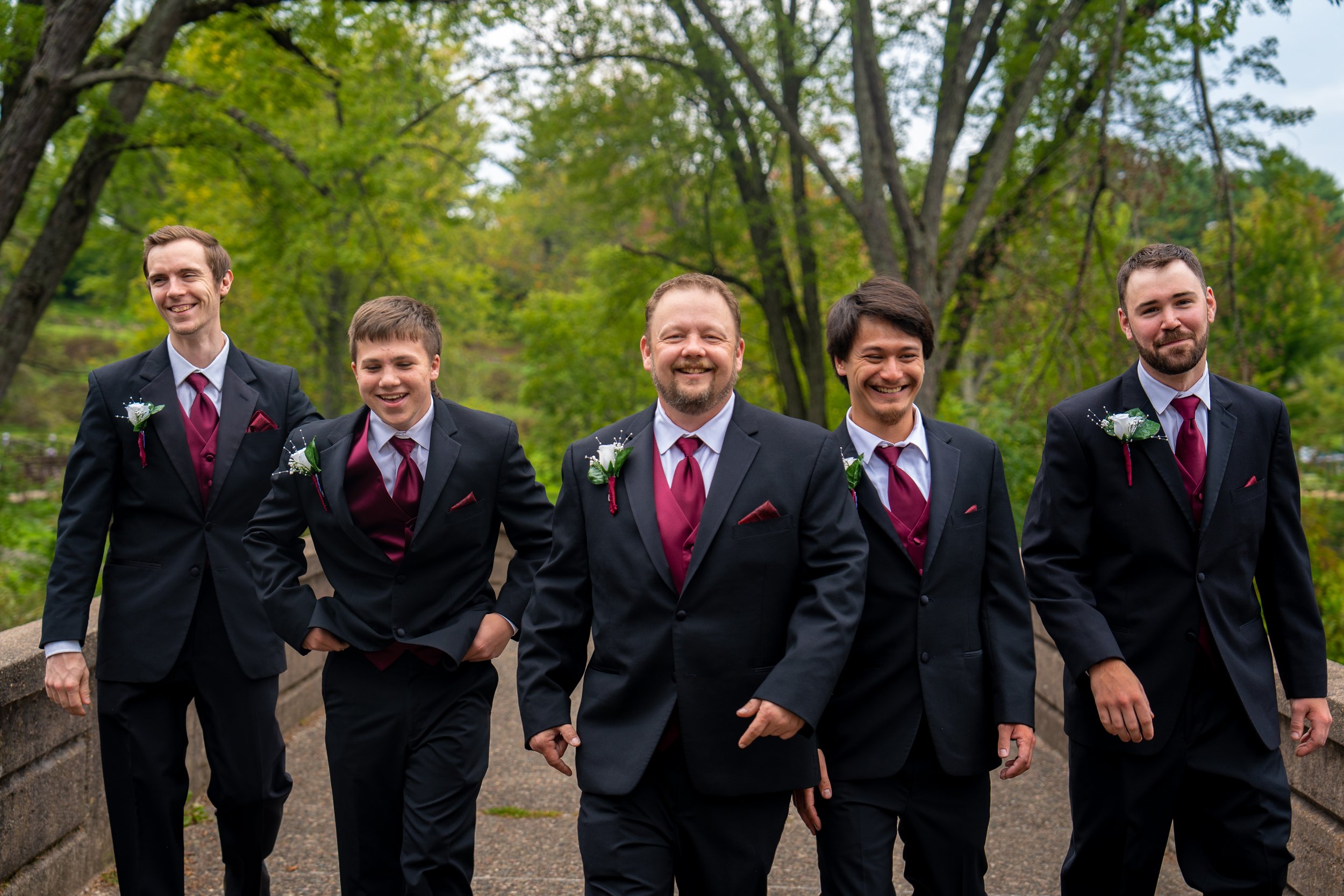 Group of five men in suits walking outdoors in a park-like setting with trees, smiling and enjoying themselves on a cloudy day.