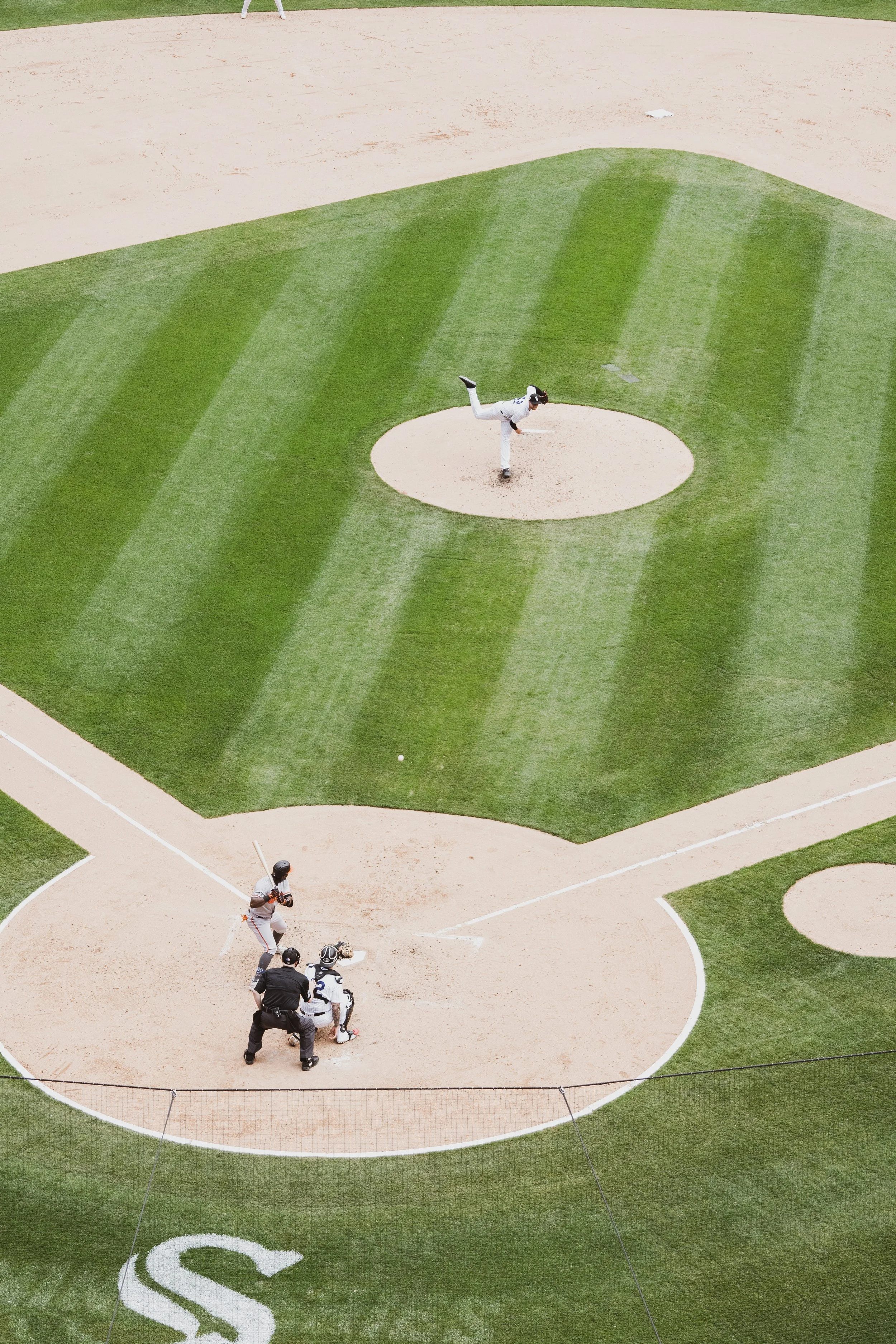 An aerial view of a baseball field shows a pitcher in white uniform throwing a pitch, a batter in gray uniform preparing to swing, an umpire behind the batter, and a catcher in black gear ready to catch the ball. The field has green grass with stripe