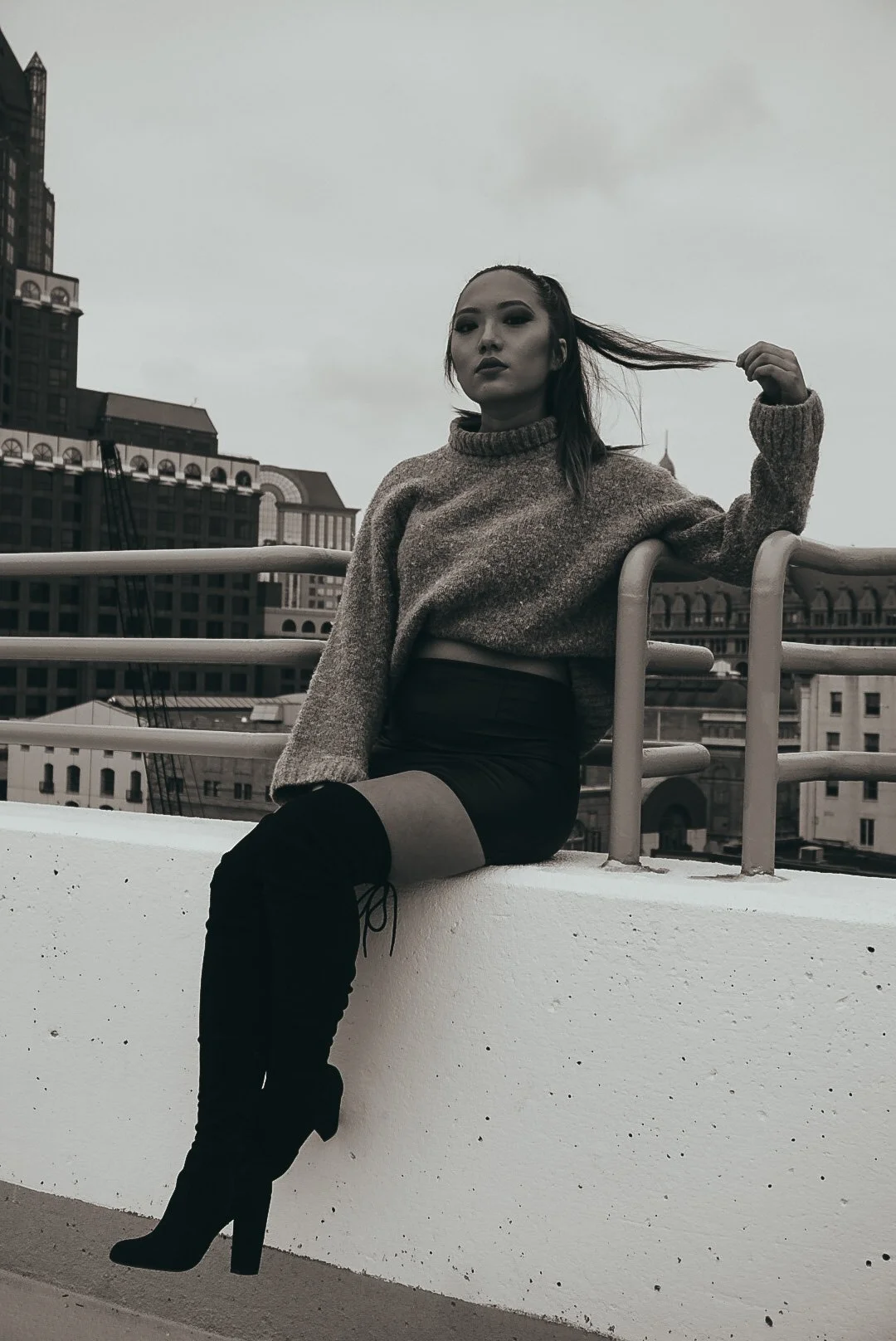 A woman sitting on a white ledge with an urban cityscape background, wearing a long-sleeve sweater and thigh-high black boots, looking serious in a black-and-white photo.