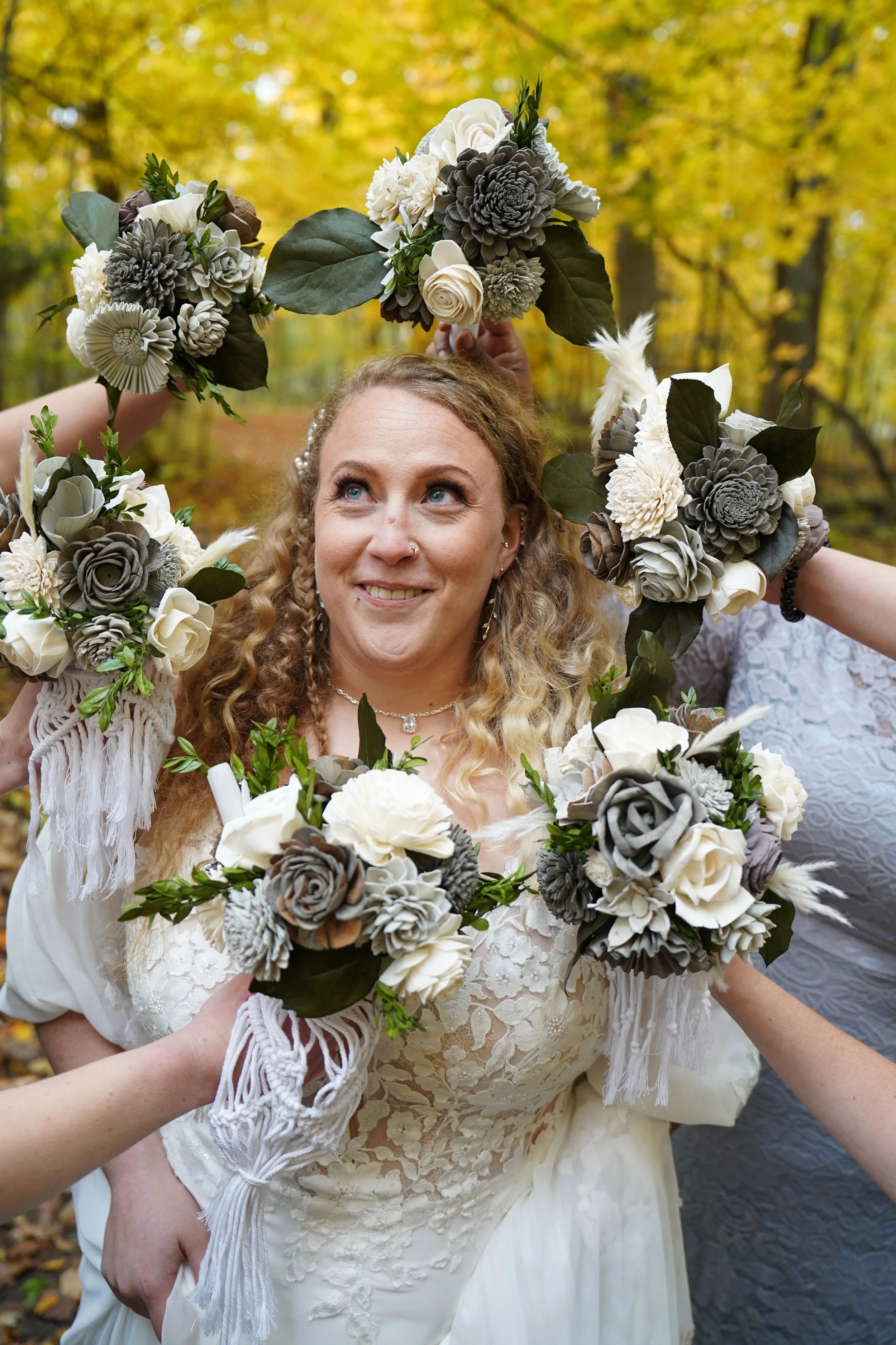 A woman with curly blonde hair and a nose piercing, wearing a white lace dress, surrounded by others holding floral wreaths in an outdoor autumn setting.