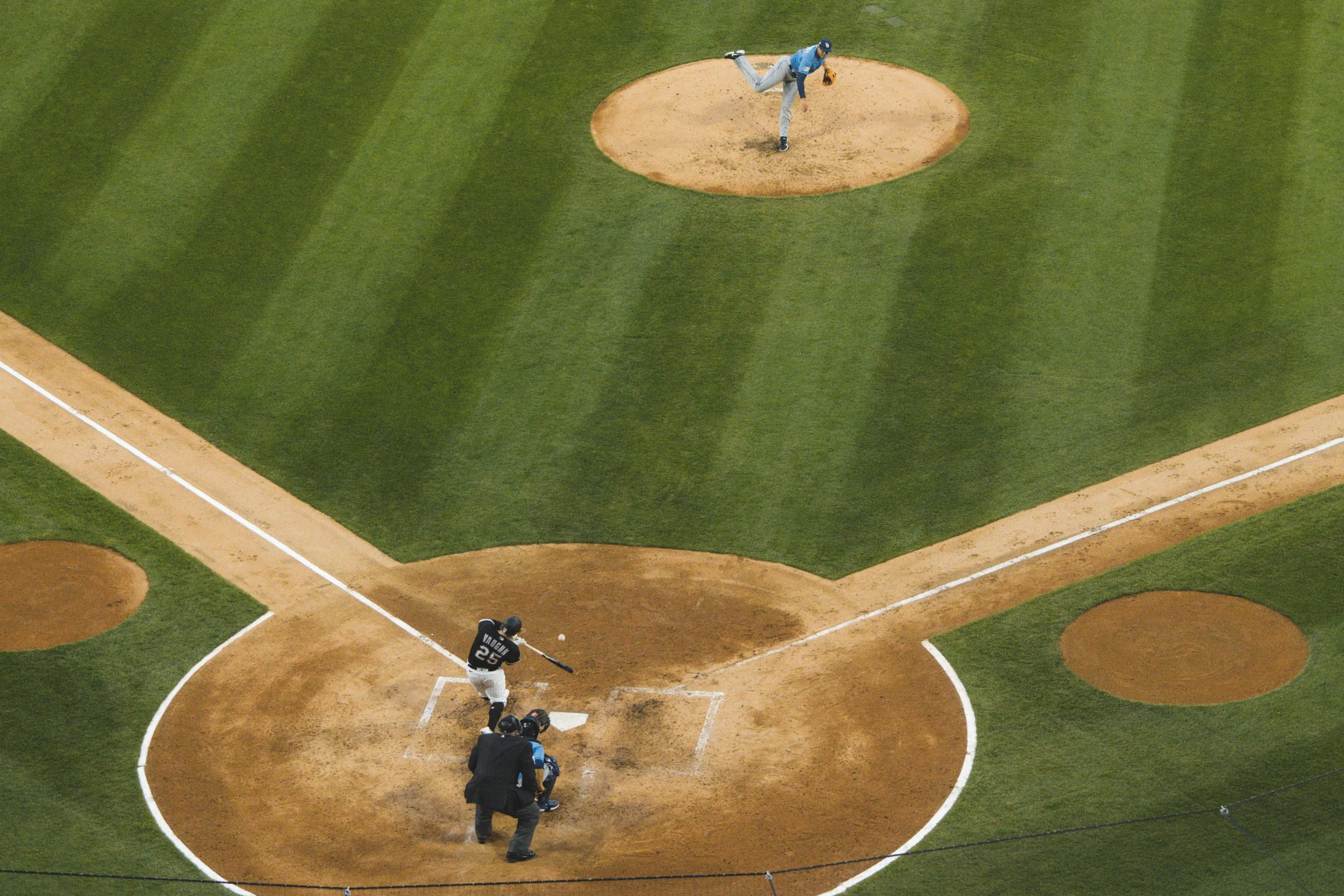 A baseball game in progress, showing a batter at home plate preparing to hit, a pitcher on the mound in the process of pitching, and a catcher behind home plate.