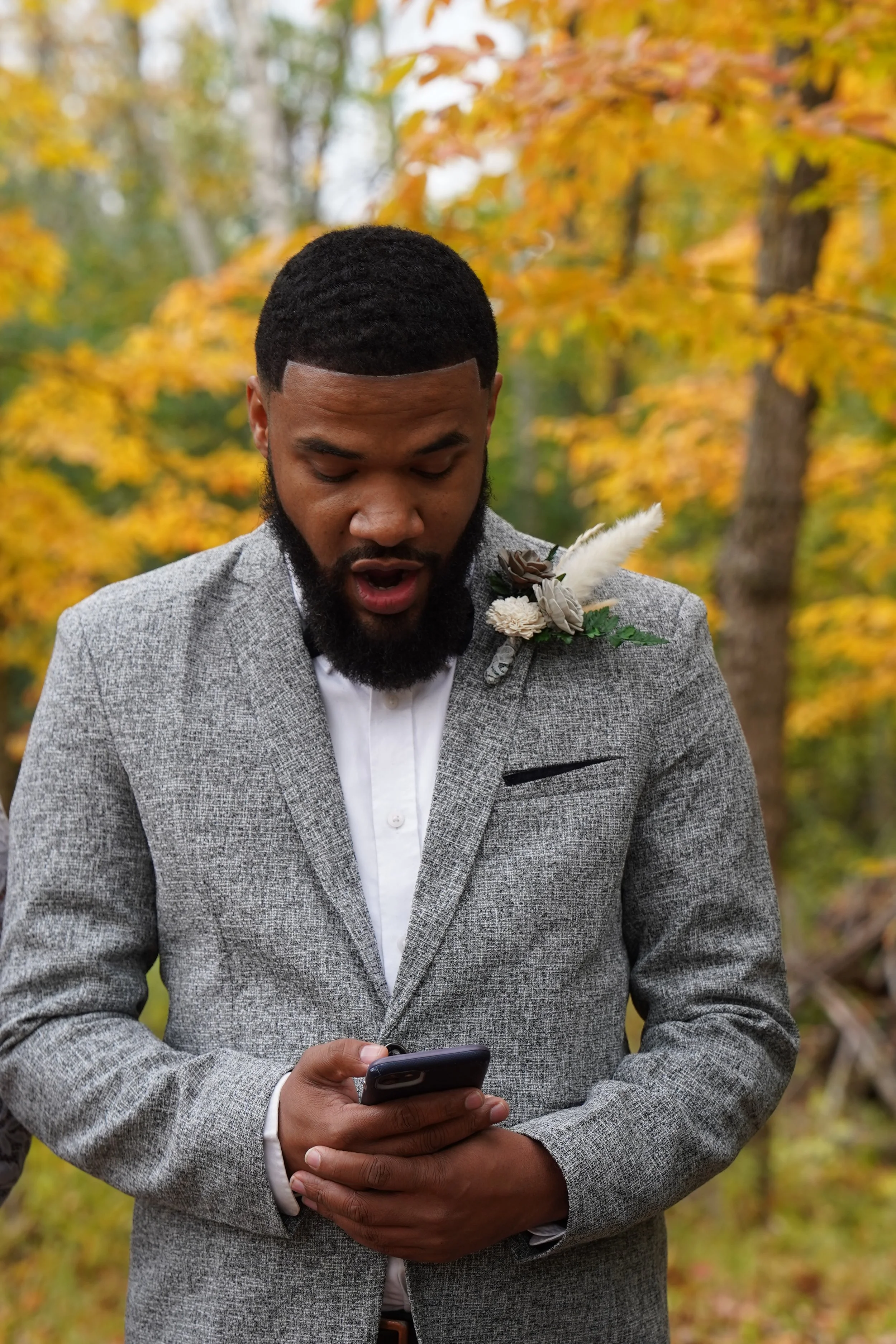 A man dressed in a gray suit with a boutonniere, standing outdoors with fall foliage in the background, looking at his smartphone with a surprised or interested expression.