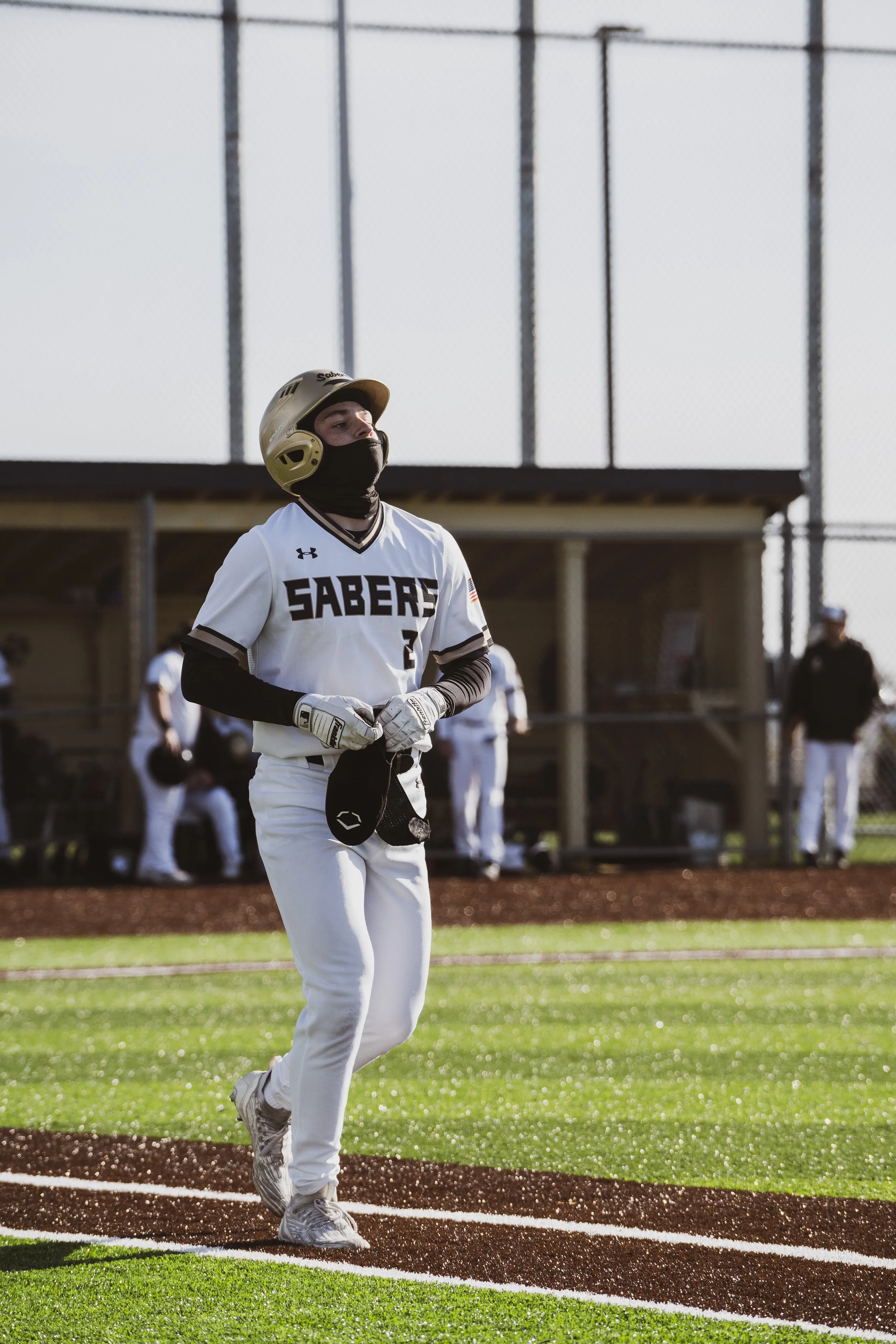A baseball player wearing a white uniform with 'SABERS' on the front, a gold helmet, black face covering, and batting gloves, standing on the field near home plate during a game.
