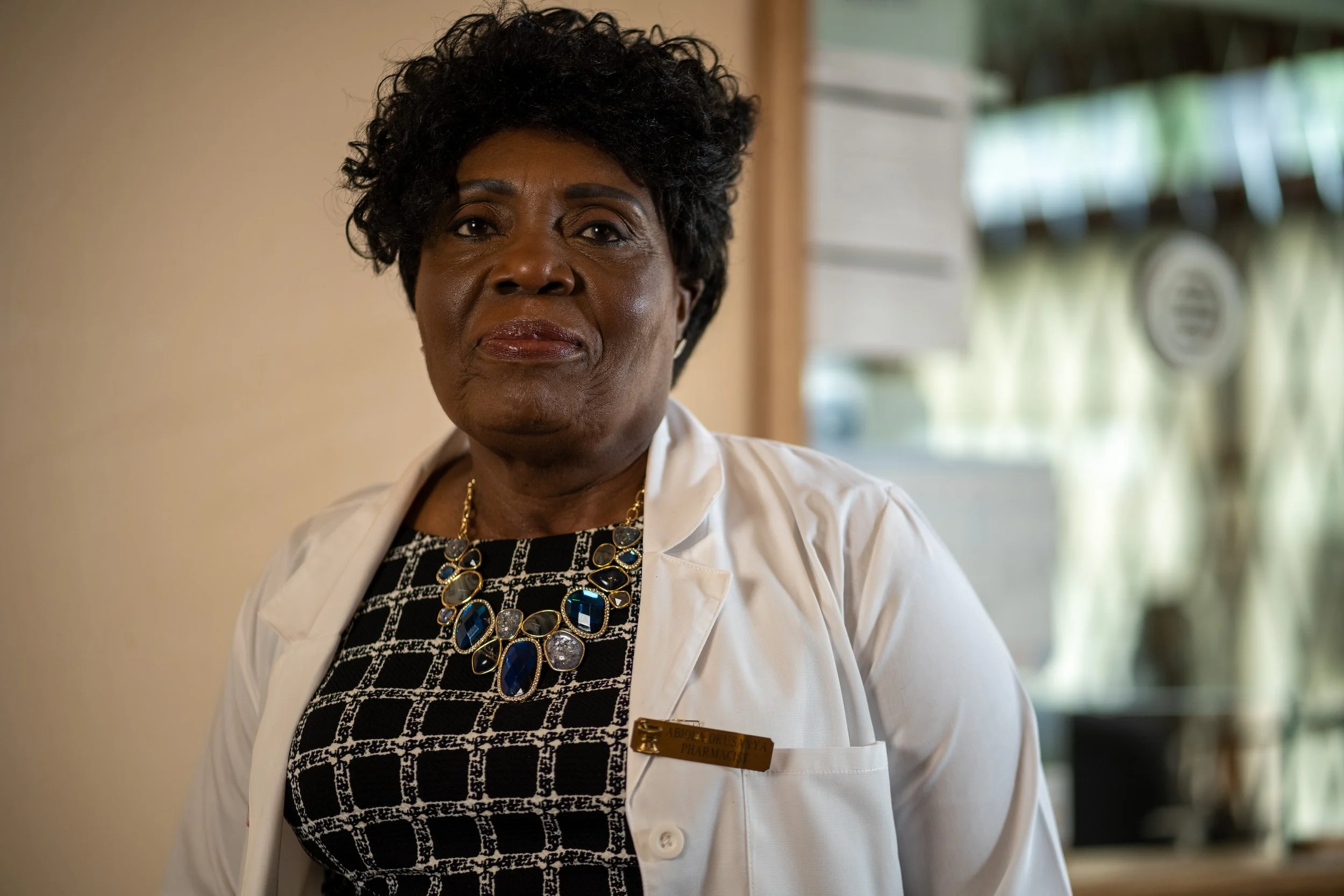 A woman wearing a white coat and jewelry, standing indoors with a neutral expression.