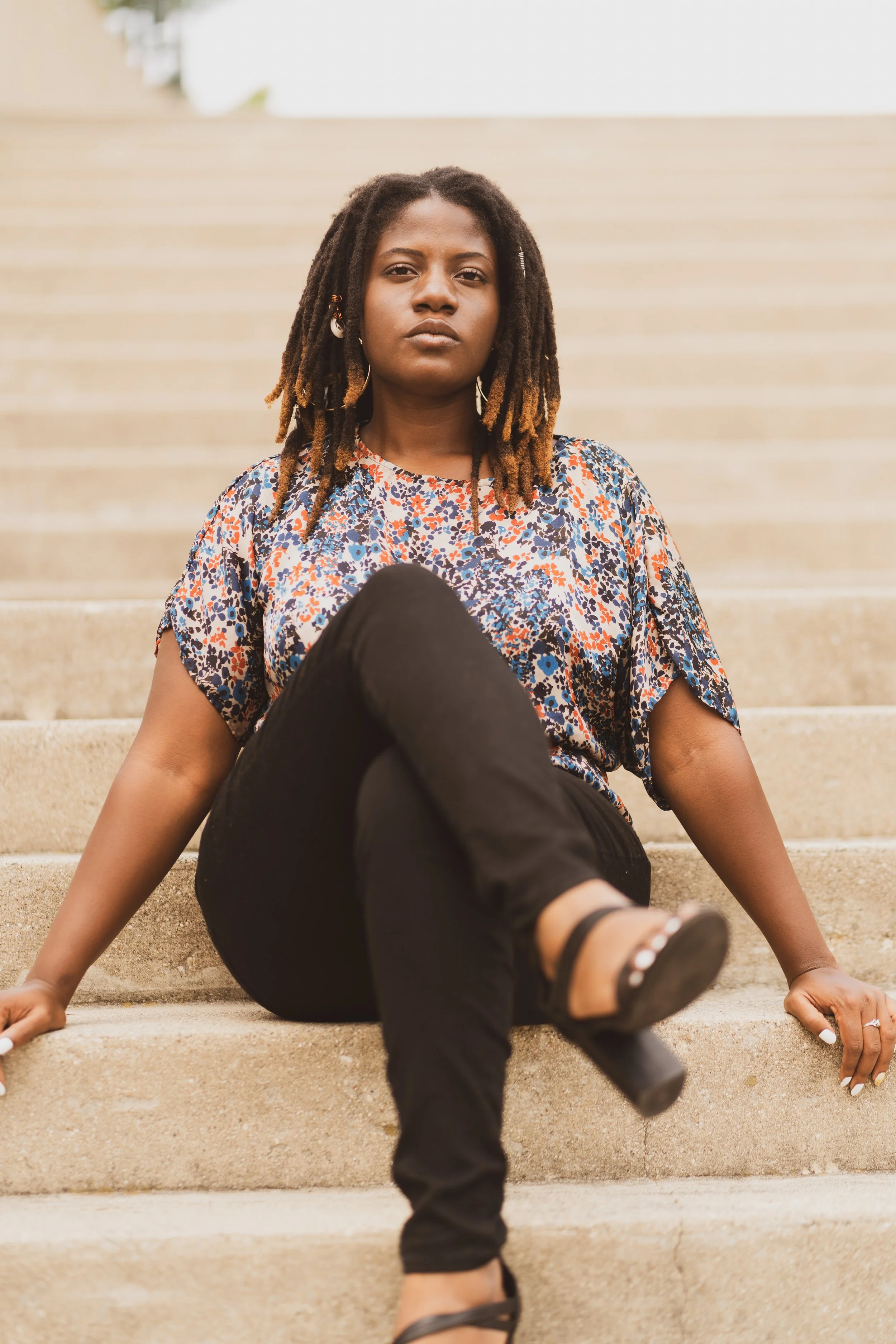 A young woman with dreadlocks sitting on outdoor concrete stairs, wearing a colorful patterned blouse and black pants, looking serious at the camera.