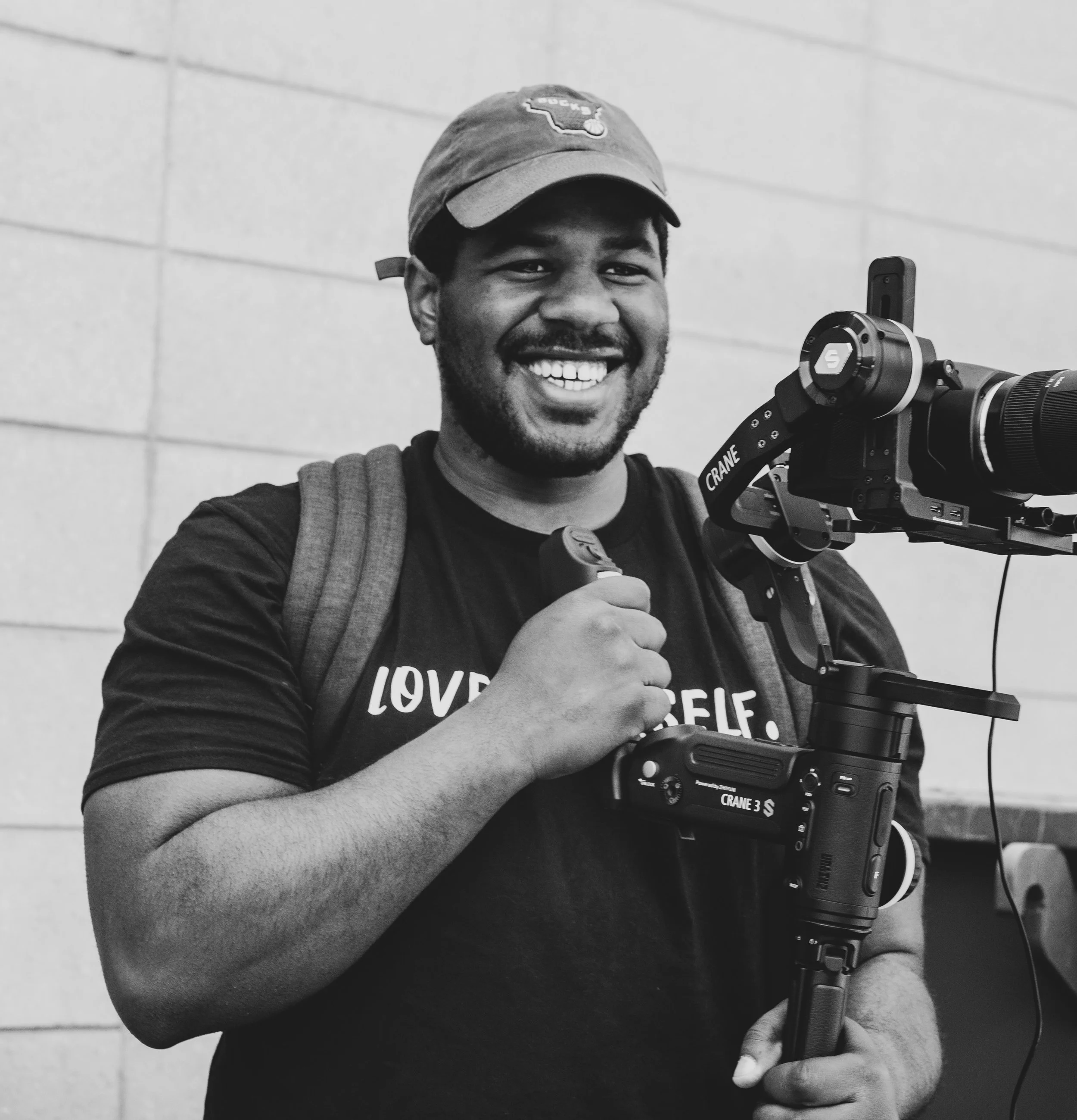 A smiling man holding a camera stabilizer and a remote control, wearing a cap and a T-shirt, standing against a plain wall.