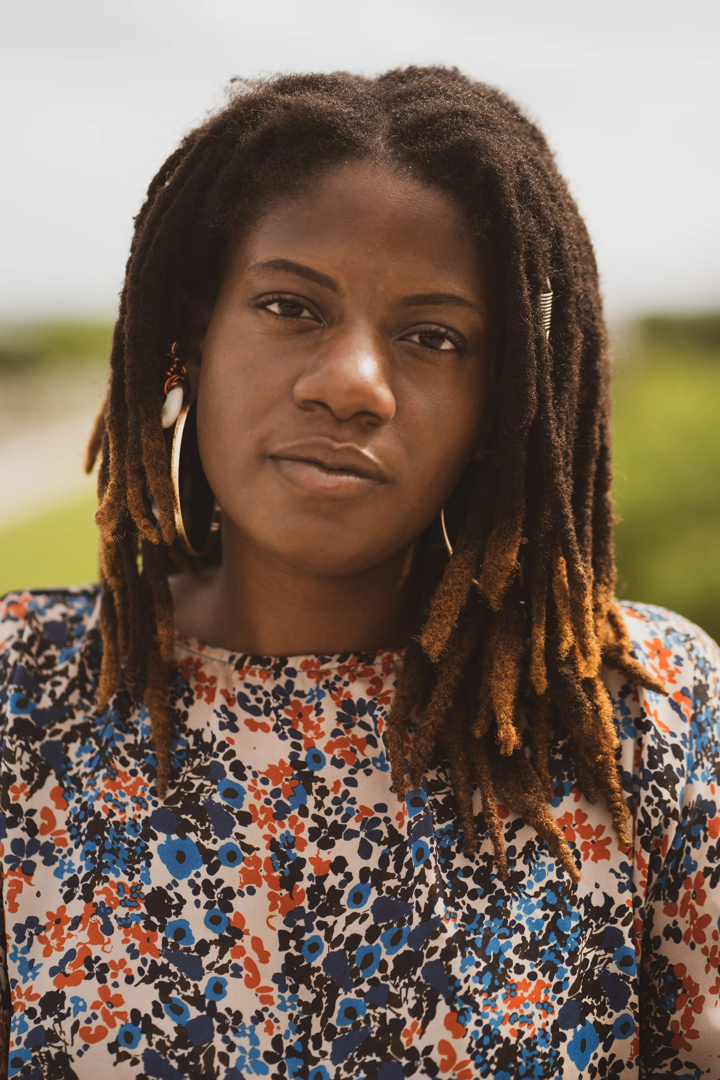Close-up of a woman with dark skin and dreadlocks wearing large hoop earrings and a floral dress, outdoors with a blurred green background.