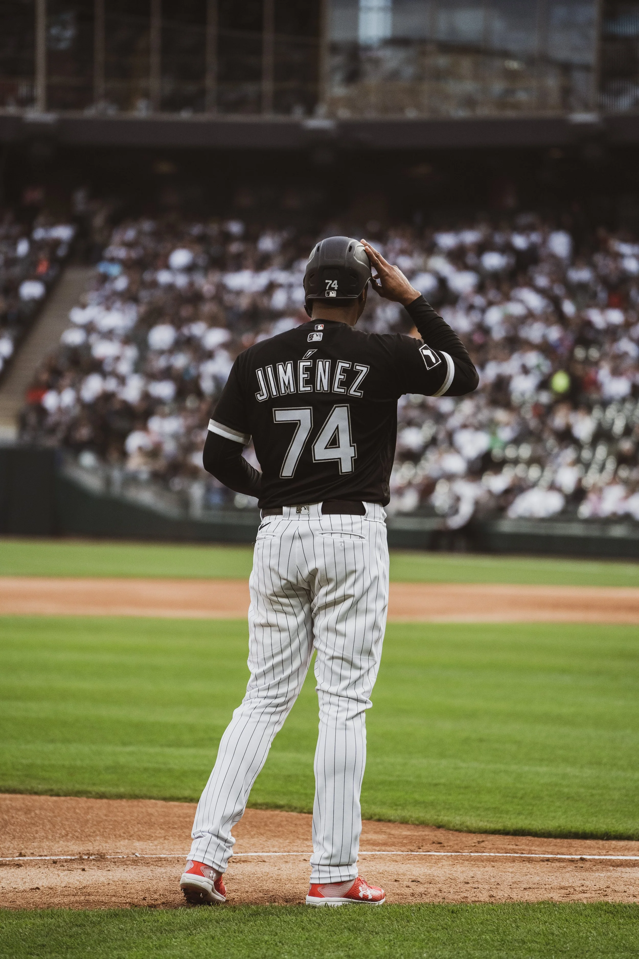 A baseball player with the name Jiménez and the number 74 on his black jersey is standing on the baseball field, holding a helmet to his head, with a large crowd of spectators in the background.