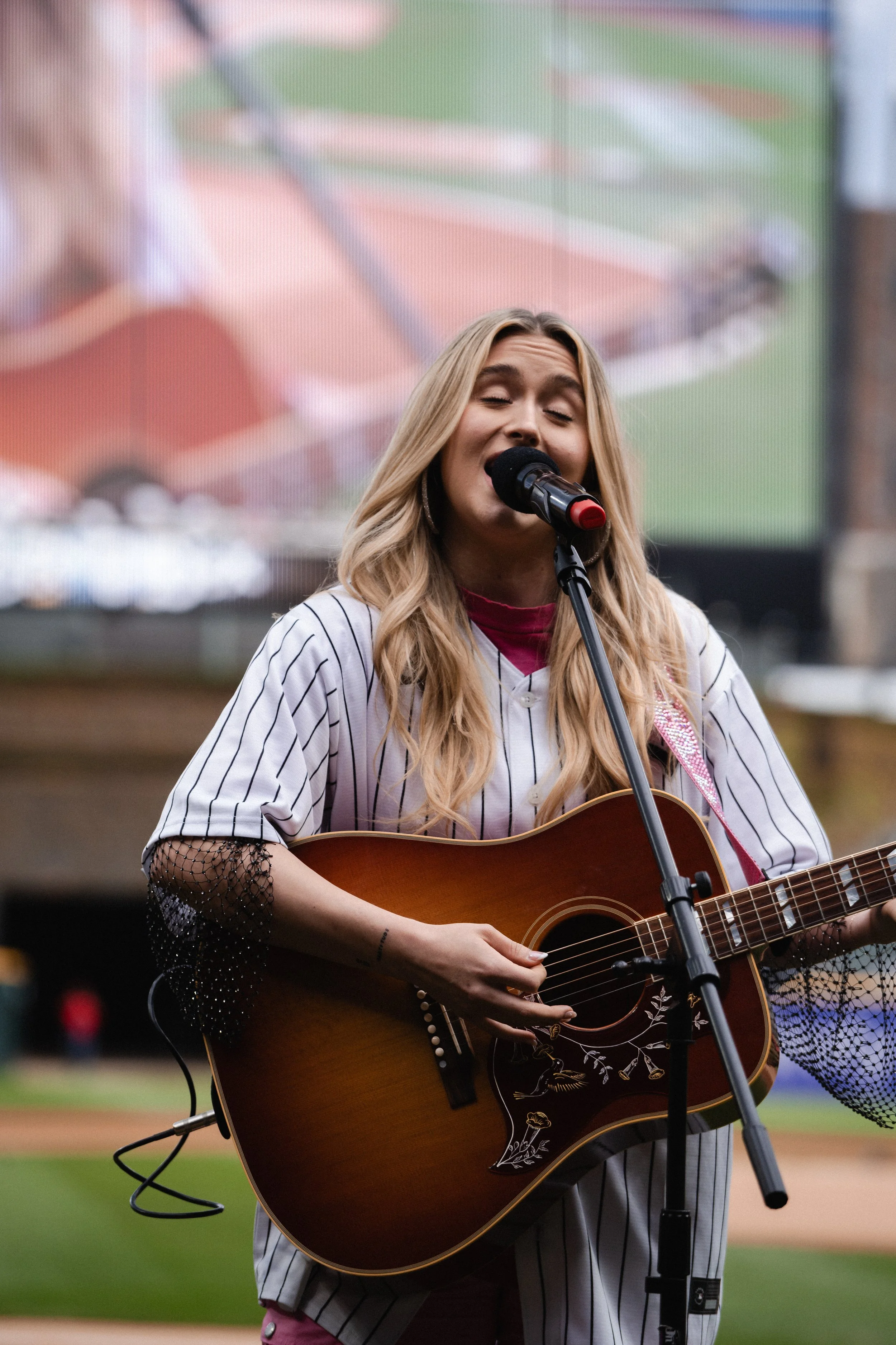 A woman with long blond hair singing into a microphone and playing an acoustic guitar on a baseball field.