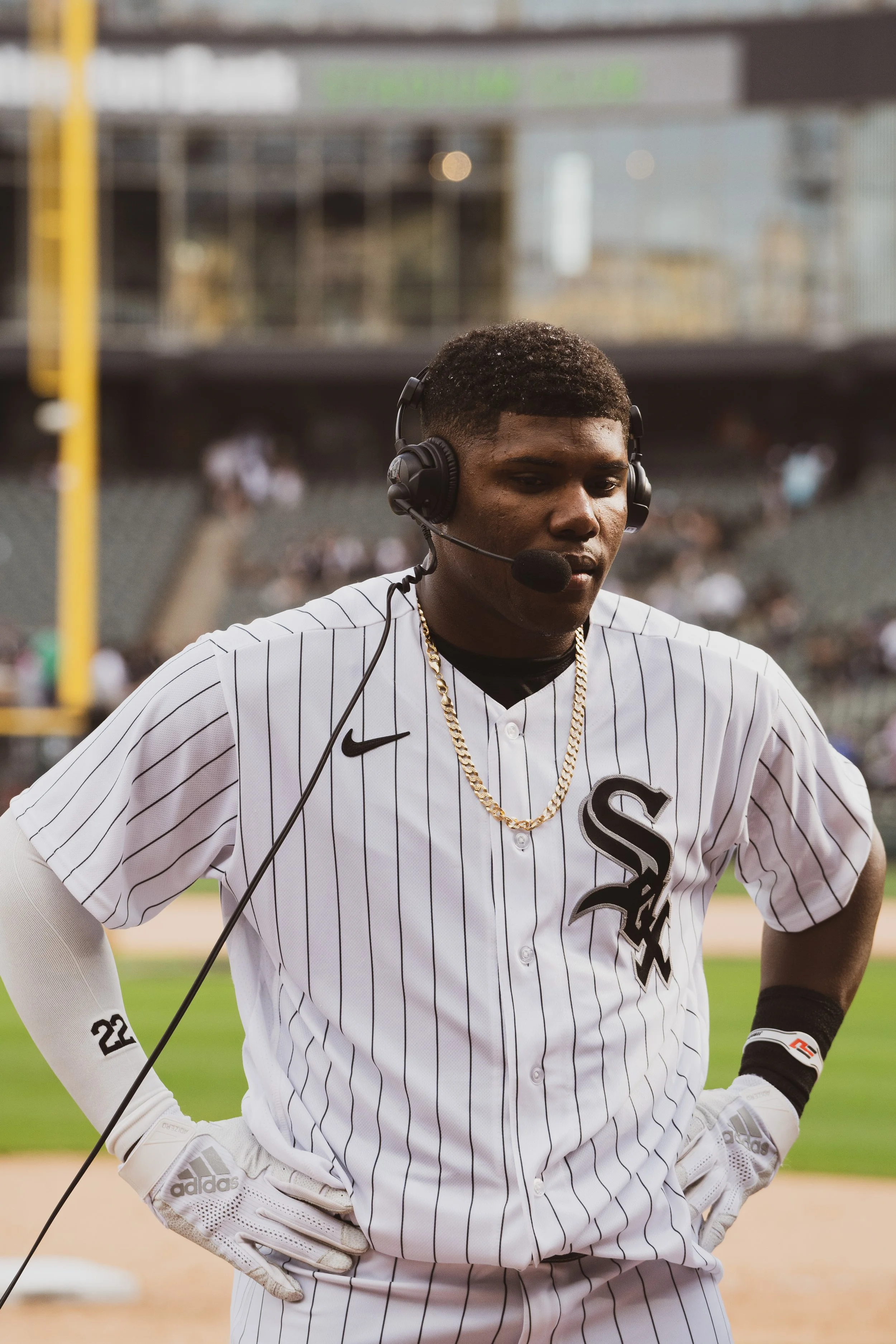 A baseball player wearing a white and black striped Chicago White Sox jersey with the number 22, standing on the field with his hands on his hips, wearing a headset and gloves, during a game at a stadium.