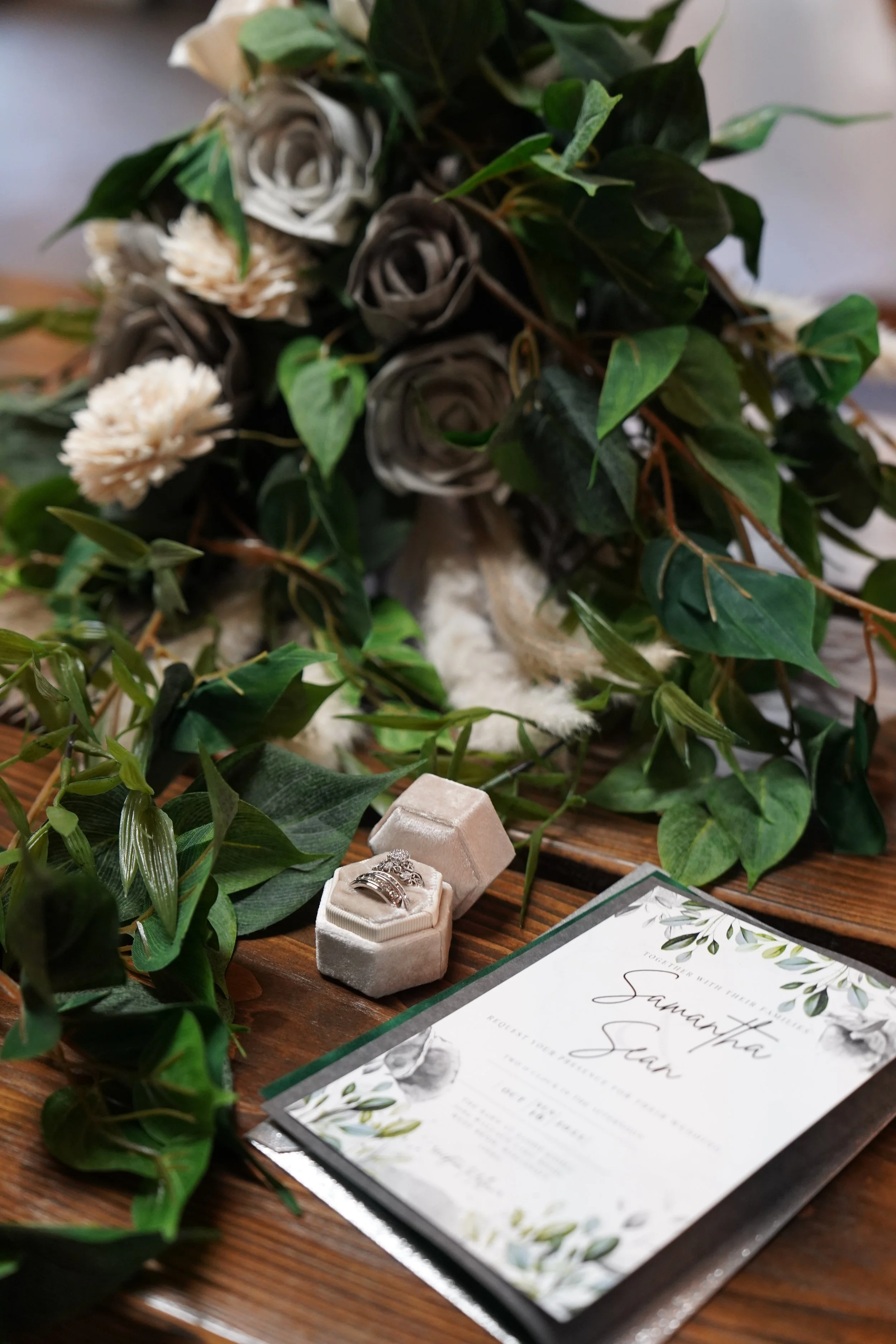 A wedding or special event table setup with a bouquet of gray and white roses, a pair of rings in a velvet box, and an invitation card with floral design and calligraphy.