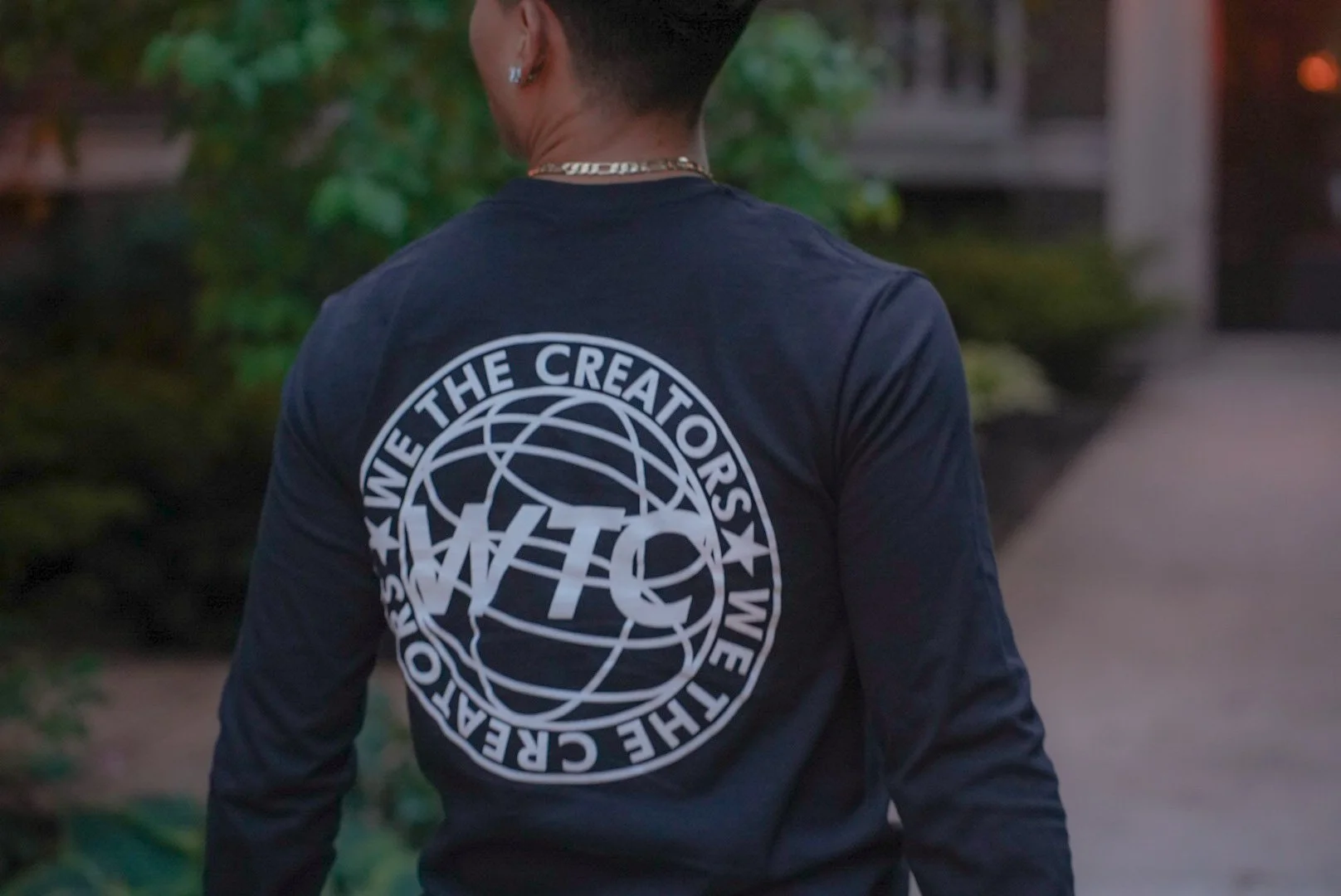 Back of a person wearing a black long sleeve shirt with a white circular logo that reads 'WIFE THE CREATORS' and 'THE CREATOR THE CREATORS' with a globe and 'WTC' in the center, walking outdoors near greenery.