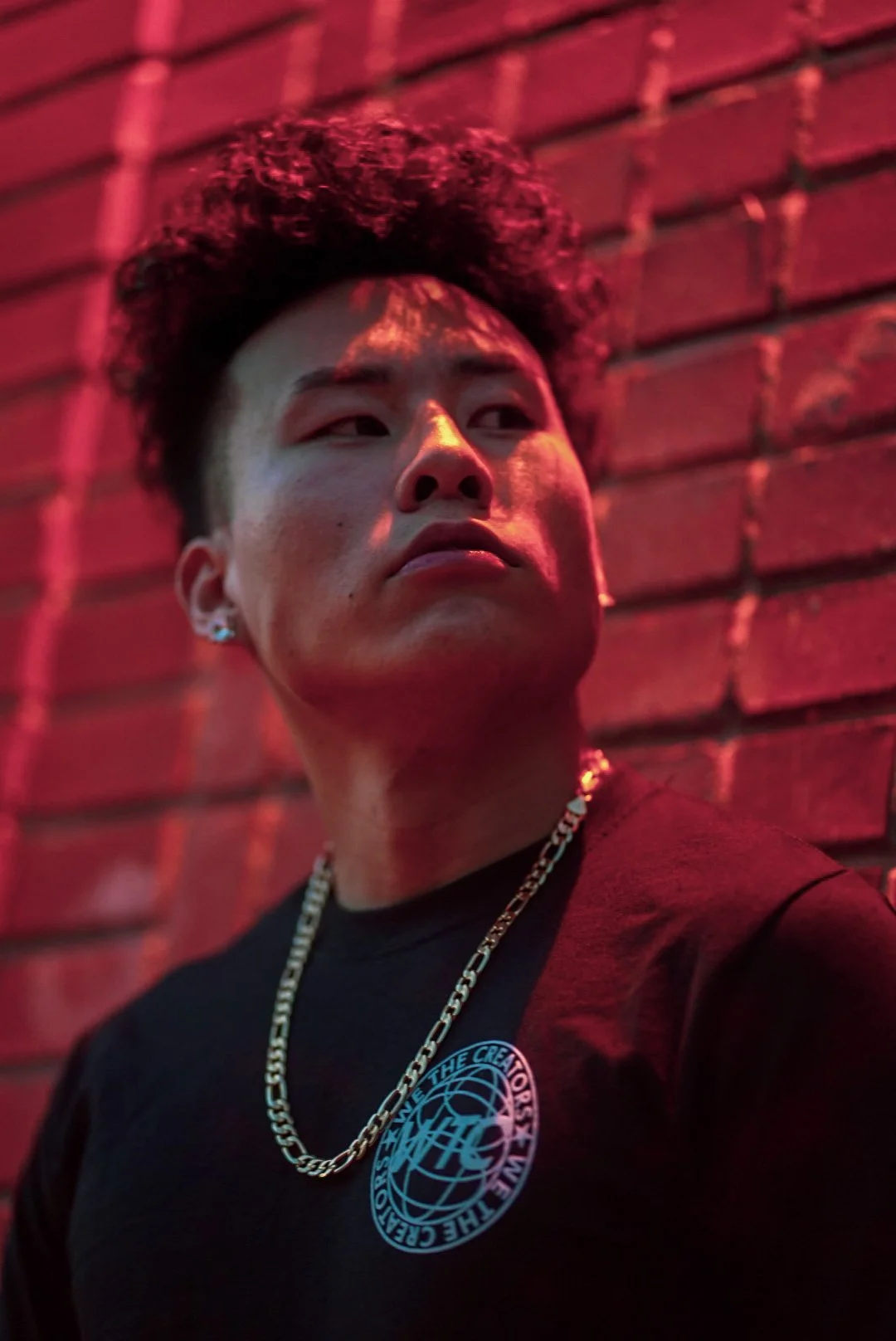 A young man with curly black hair, earrings, and a chain necklace standing against a brick wall illuminated by red and pink lighting.