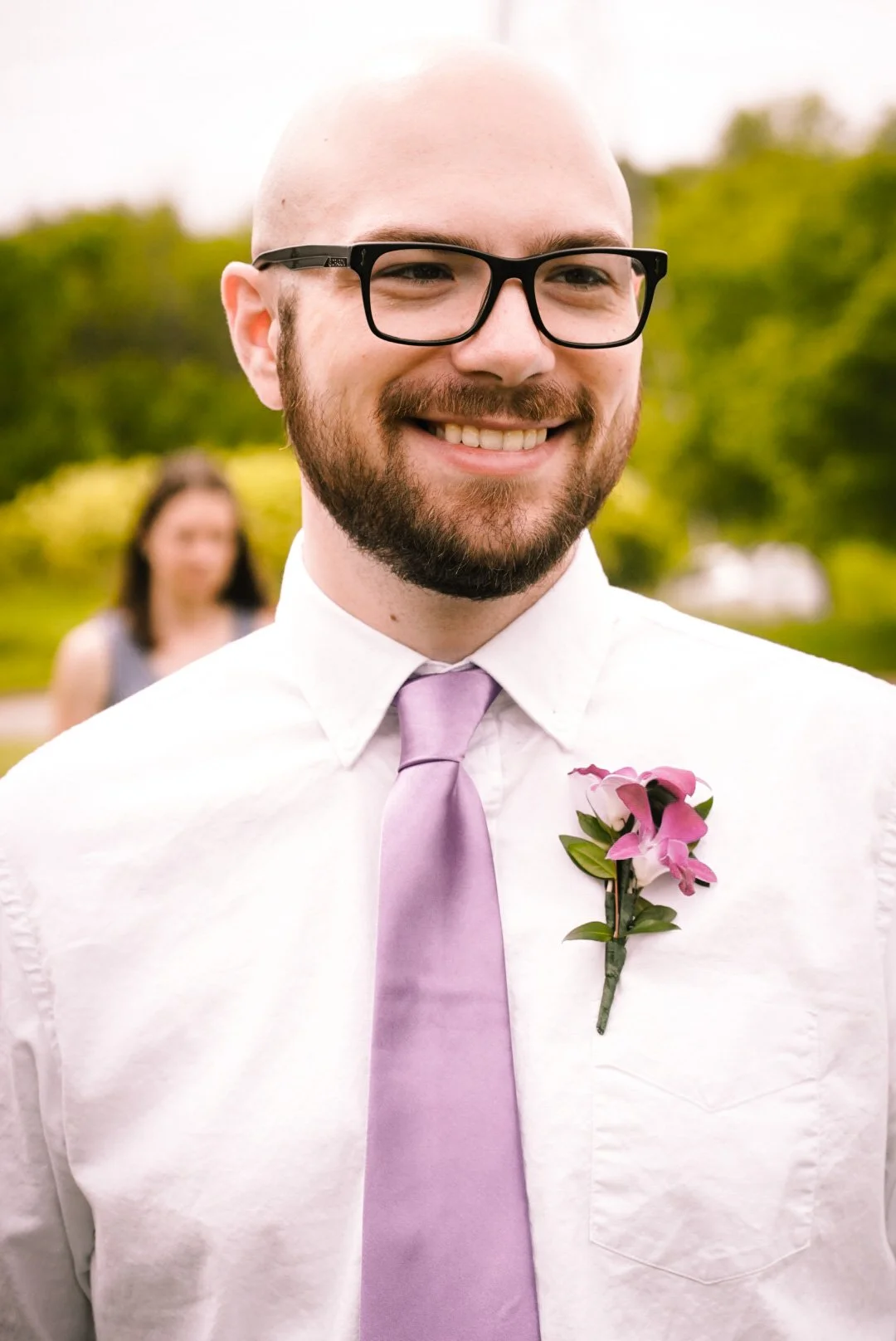 Close-up of smiling man with glasses, beard, wearing a white shirt and lavender tie with pink flower boutonniere, outdoors, blurred woman in background.
