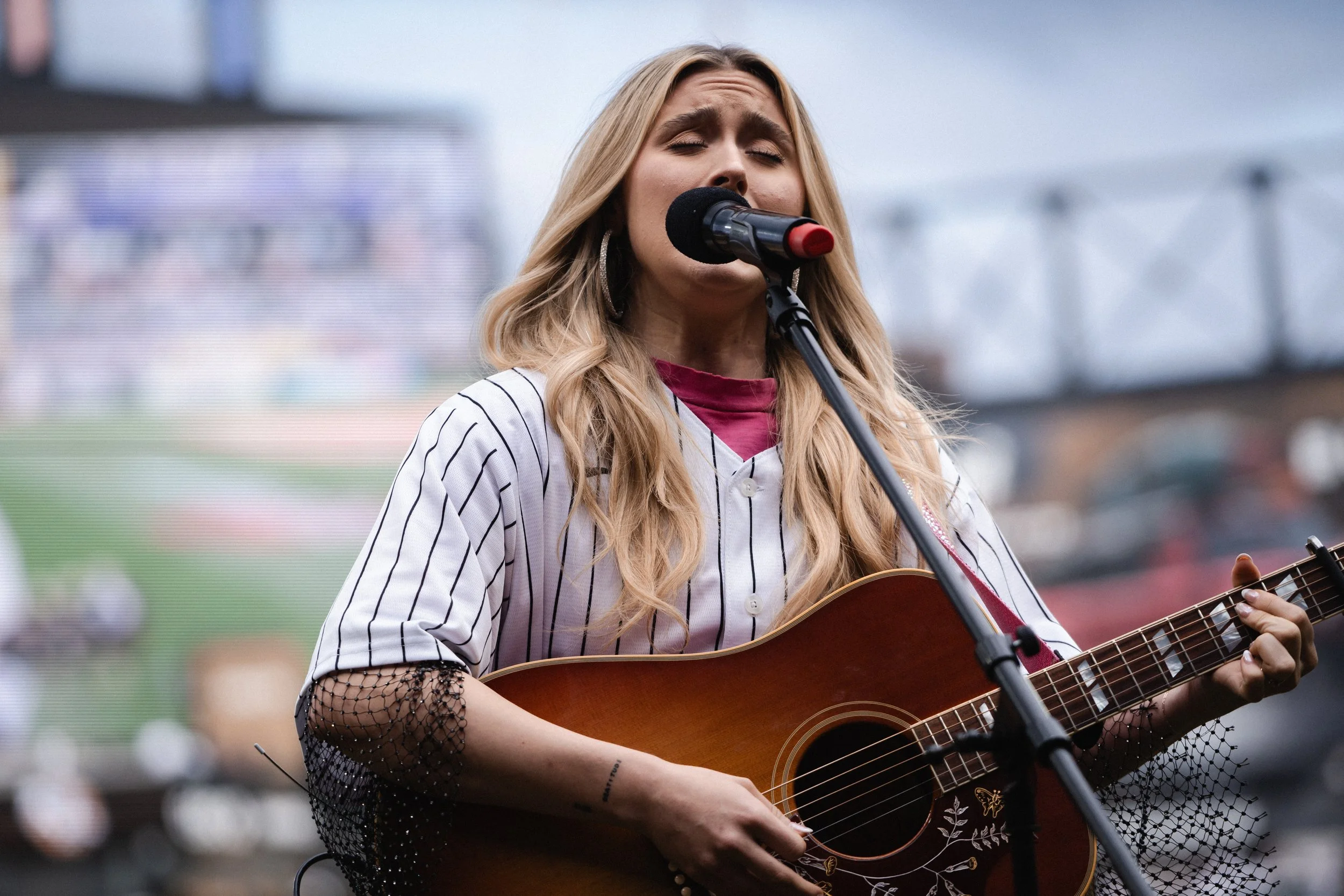 A woman with long blonde hair singing into a microphone and playing an acoustic guitar during a live outdoor performance.
