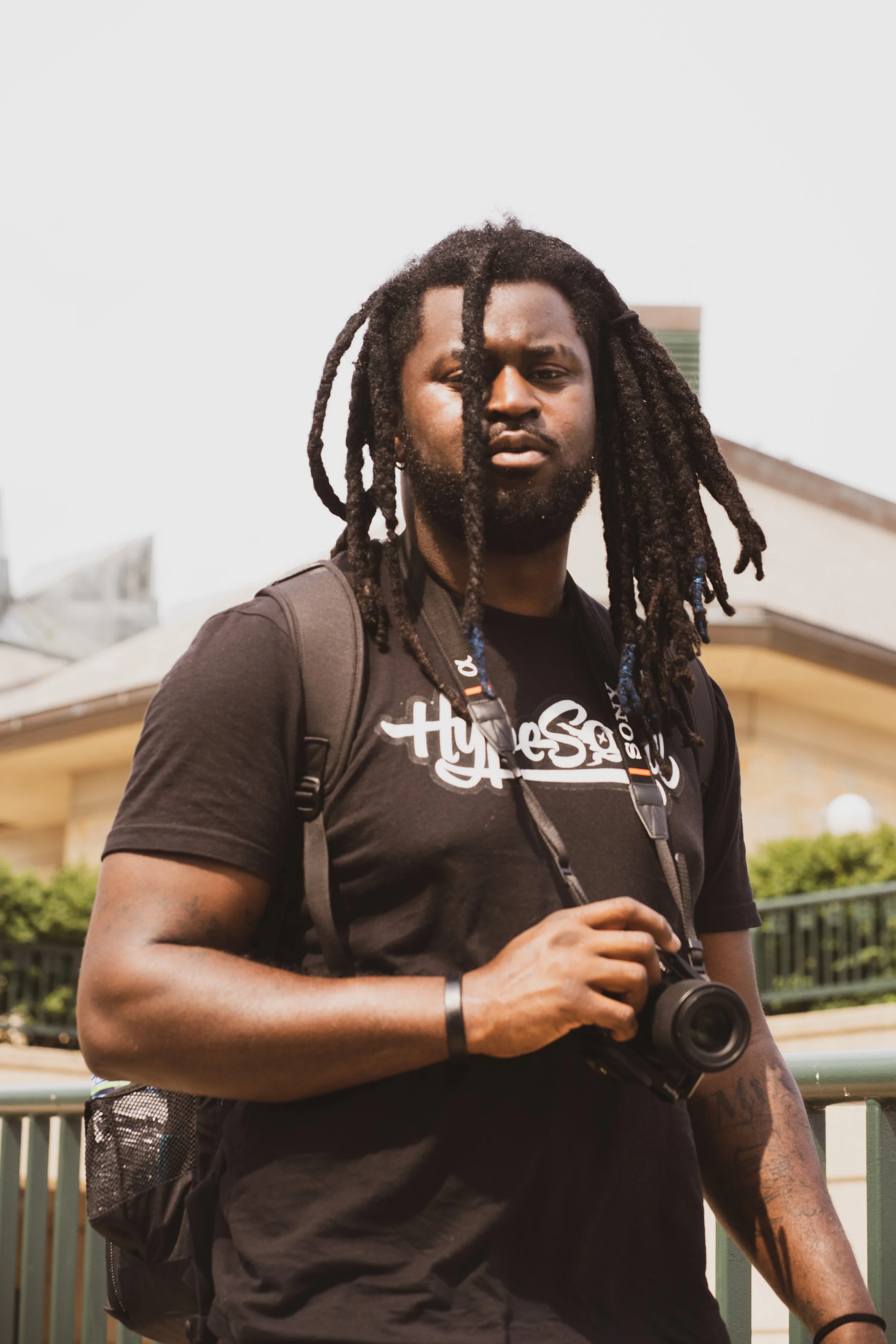 A young man with dreadlocks holding a camera, wearing a black t-shirt and backpack outdoors.
