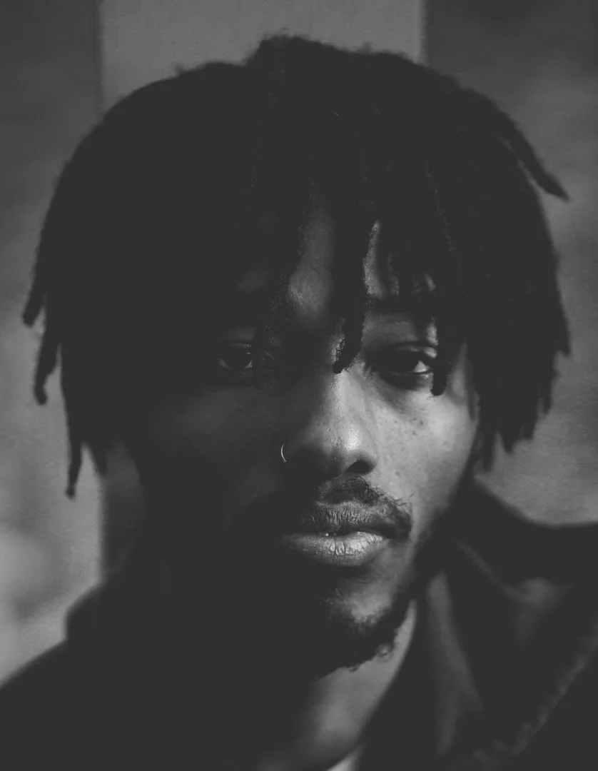 Black and white close-up portrait of a young man with dreadlocks, a nose piercing, and a serious expression.