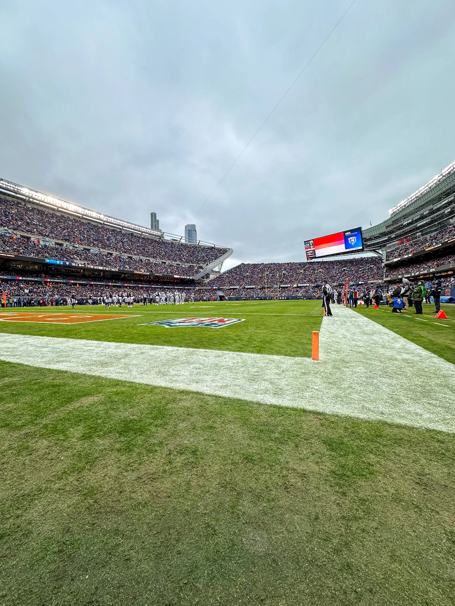 Wide view of a football stadium with a game in progress, filled with spectators, cloudy sky overhead, field markings visible, and players near the end zone.