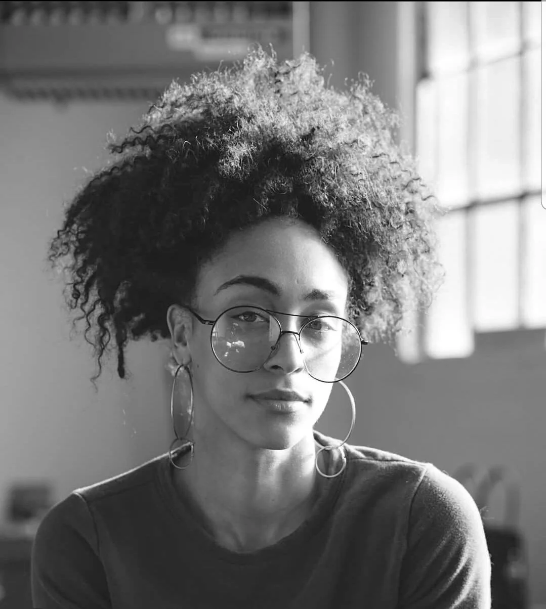 Black and white photo of a woman with curly hair, wearing glasses and large hoop earrings, looking at the camera.