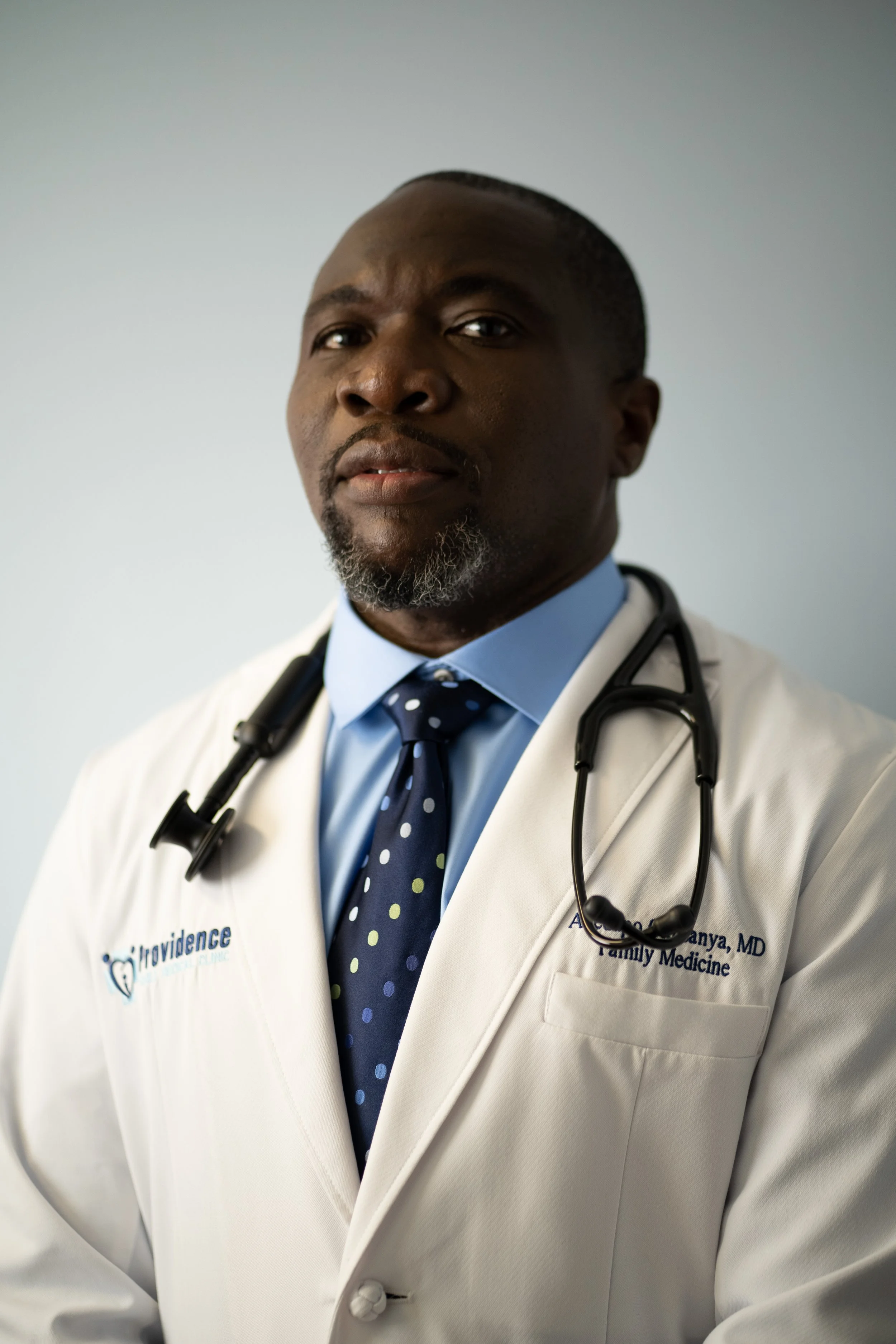 African American male doctor wearing white coat, blue dress shirt, dotted tie, with stethoscope around neck, in a professional portrait.