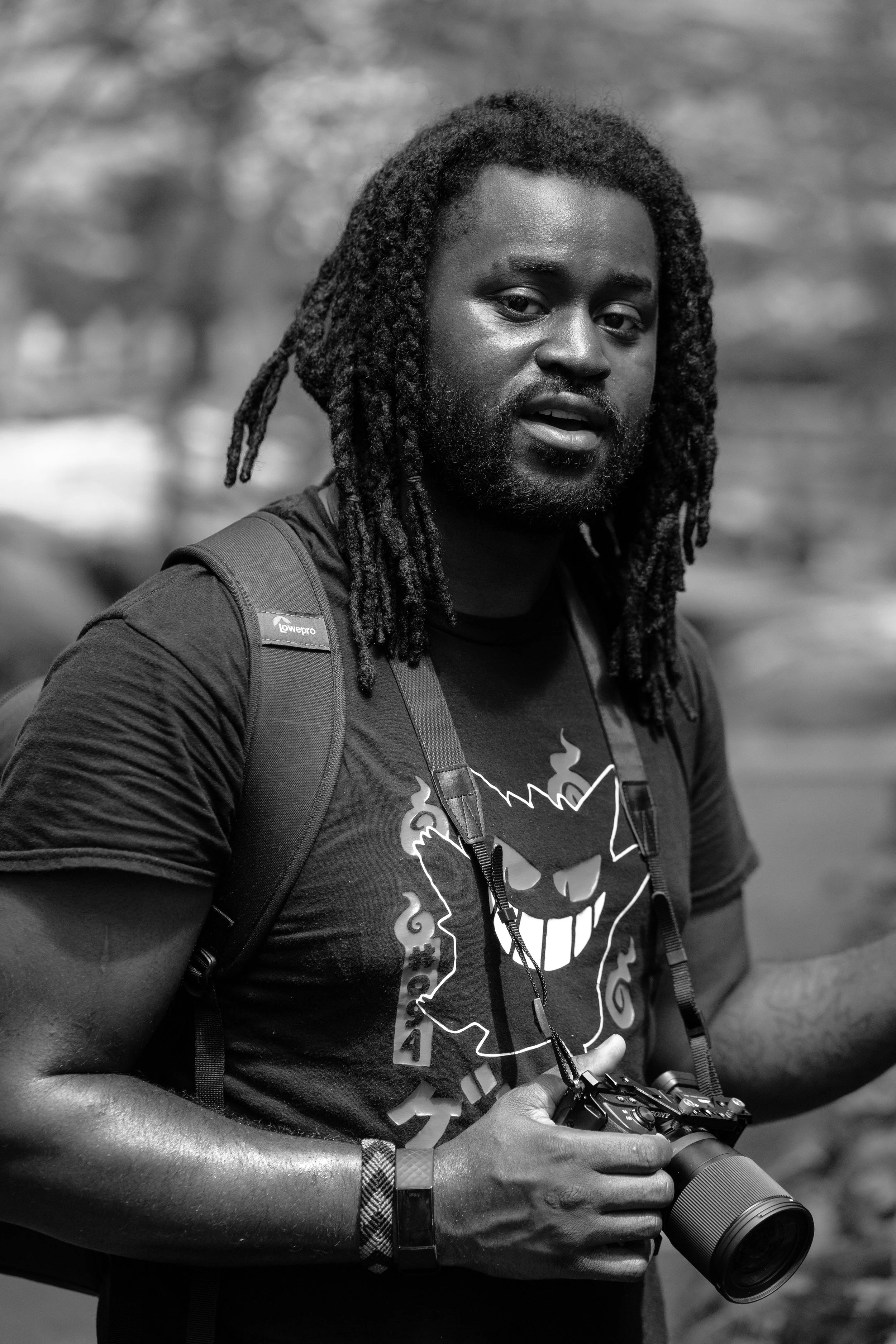 A man with dreadlocks holding a camera, wearing a graphic T-shirt with a smiling face, and a backpack, outdoors.