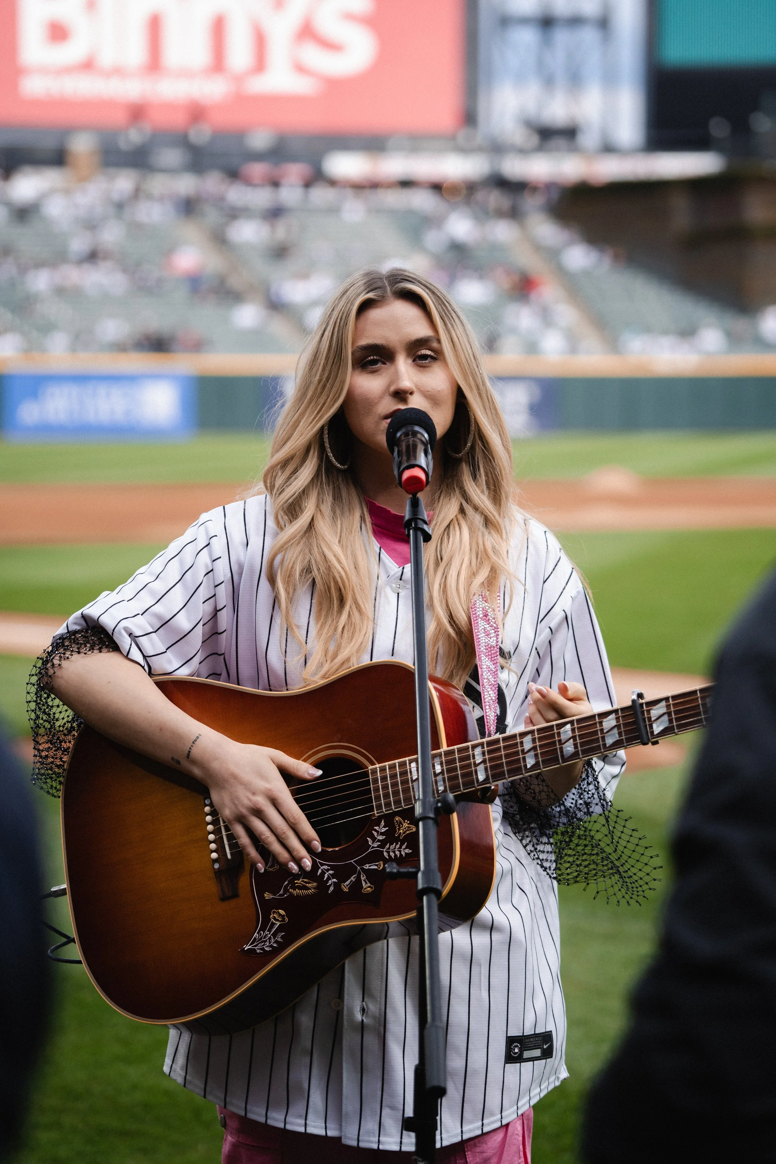 A woman with long blonde hair singing into a microphone while playing an acoustic guitar at a baseball stadium.