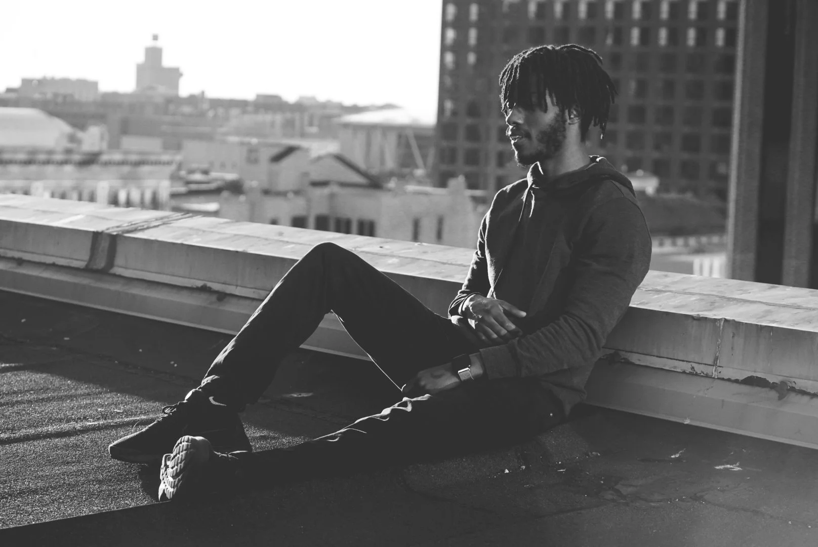 A man with dreadlocks wearing a hoodie and dark pants, sitting on a rooftop ledge in an urban cityscape, with buildings in the background, captured in black and white.