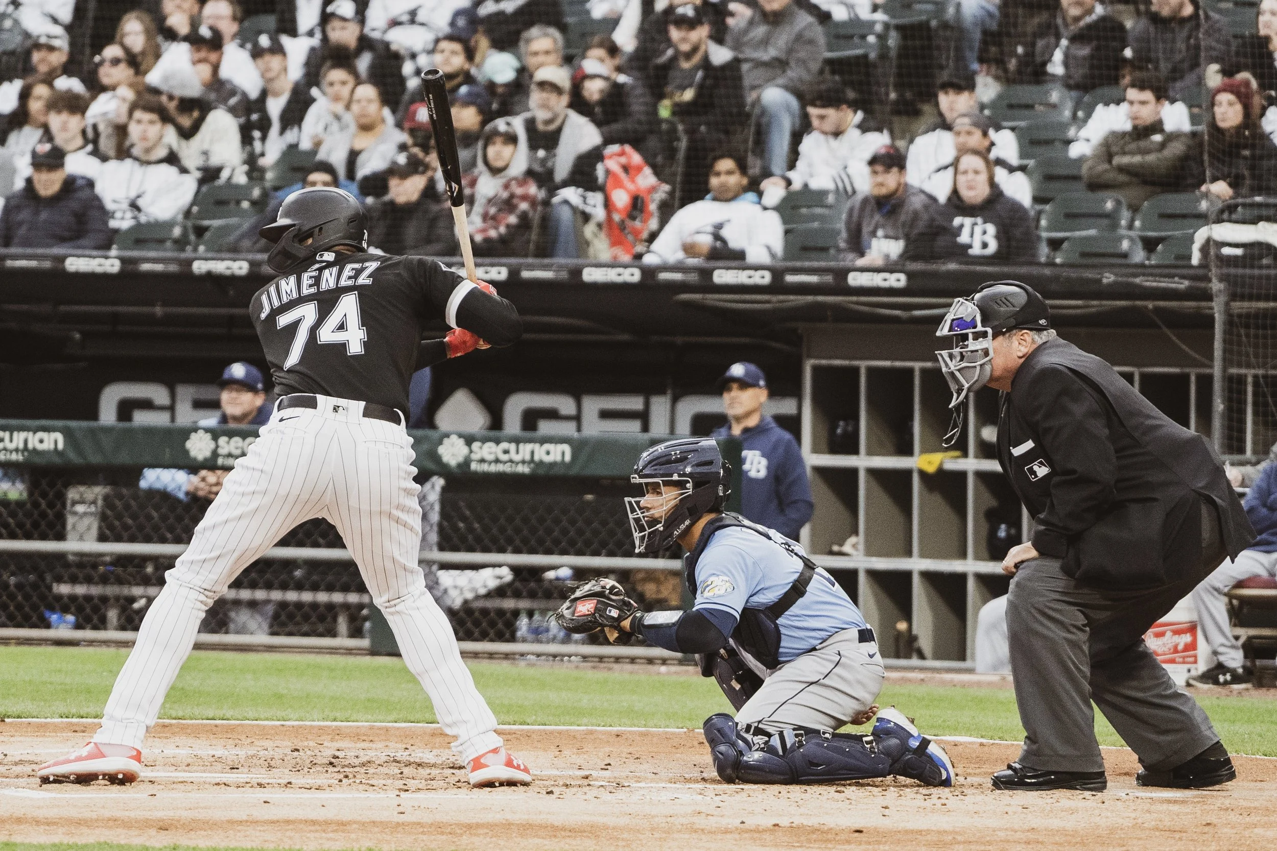 Baseball player at bat with catcher and umpire behind home plate, and spectators in the stands.