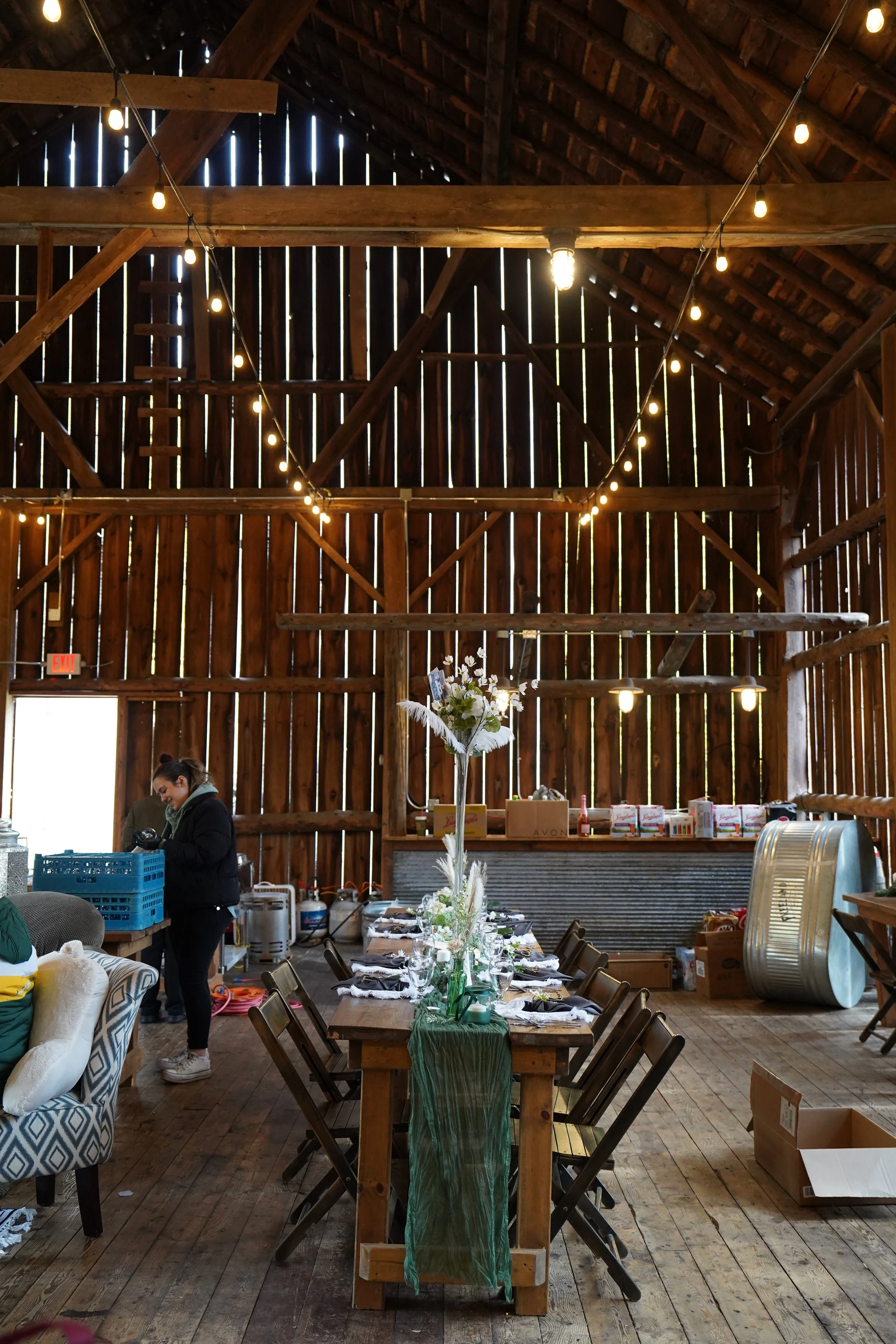 Interior of a rustic wooden barn with a long dining table set with black and white napkins, flowers, and tableware. String lights hang from the ceiling. A woman is standing near a blue crate, and boxes and supplies are on shelves and the floor.