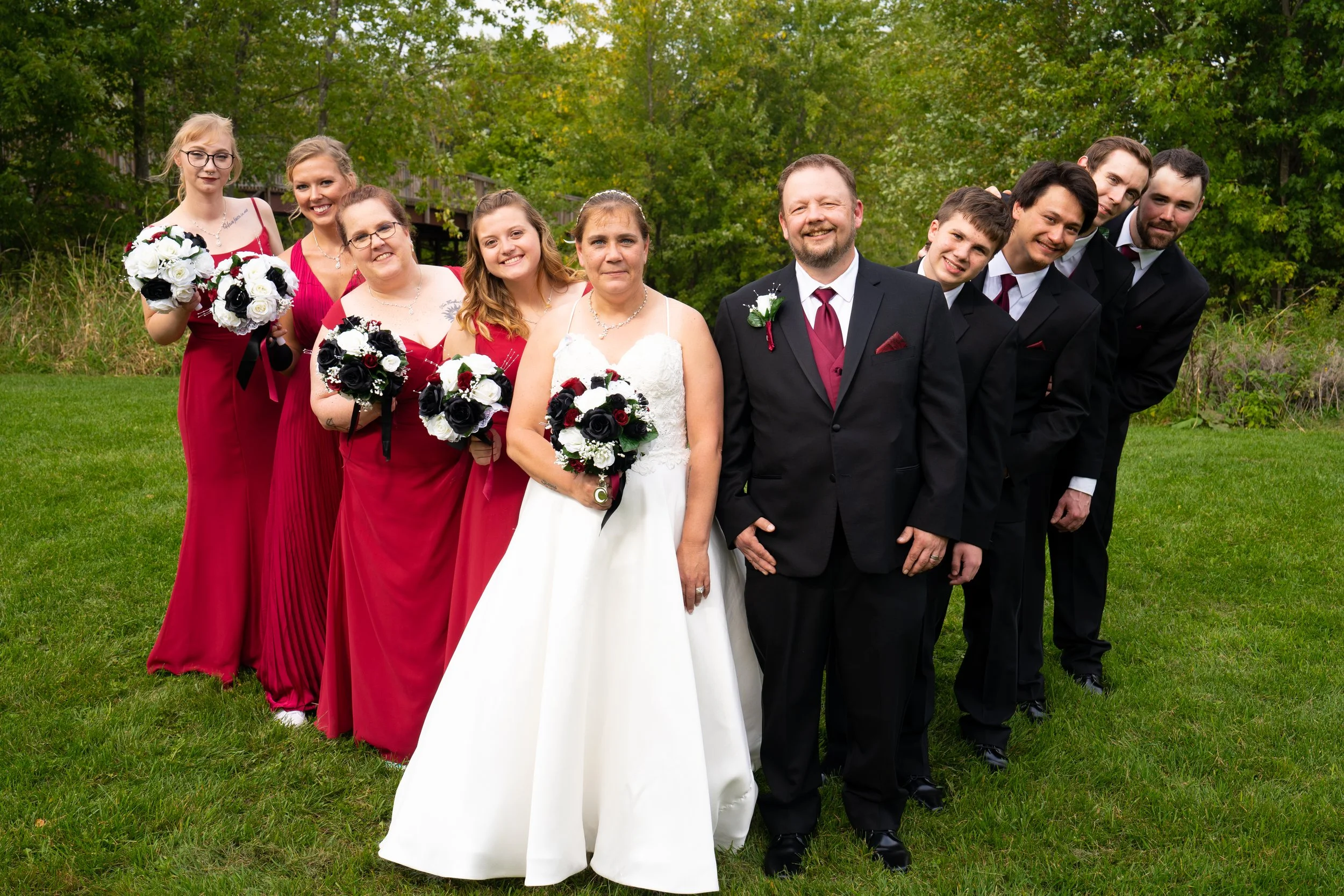 Group of wedding party members, including bride, groom, bridesmaids, and groomsmen, standing on grass outdoors surrounded by green trees, dressed in formal attire with bouquets and boutonnieres.